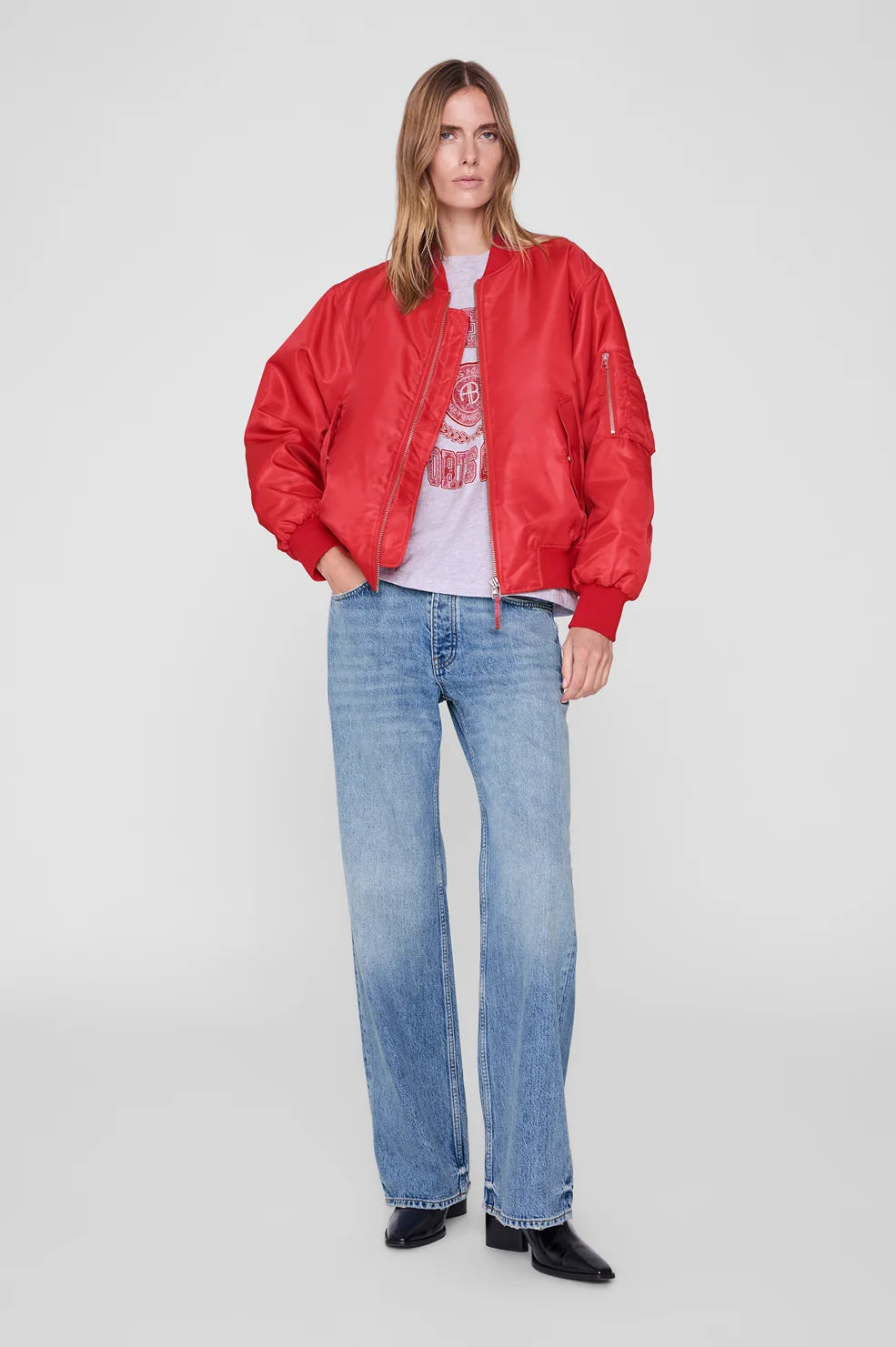 A woman modeling a bright red bomber jacket over a white graphic t-shirt and light-wash blue jeans, standing against a light gray studio background.