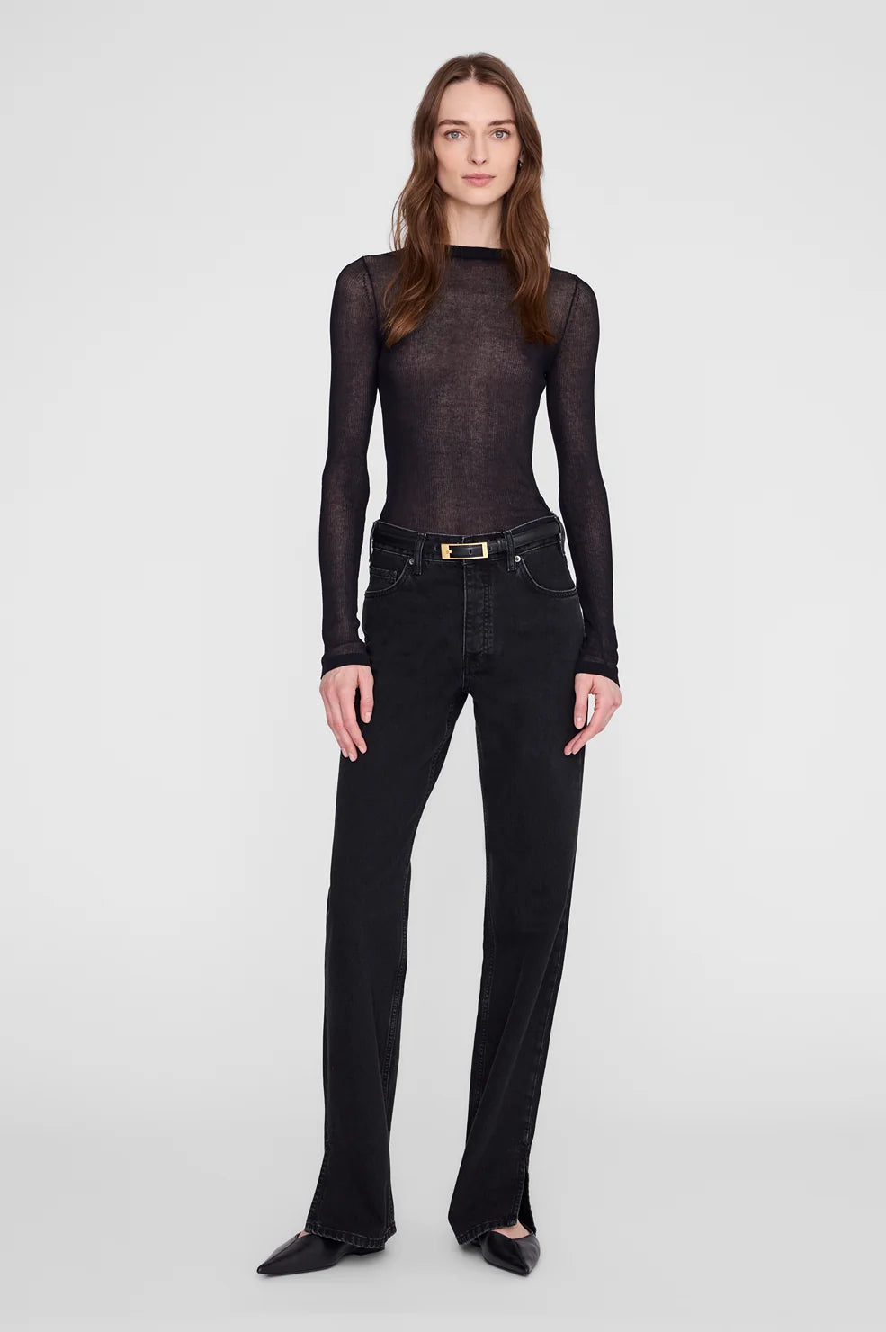 A woman modeling a sheer black long-sleeve mock-neck top paired with black jeans and black pointed flats, standing against a light gray studio background.