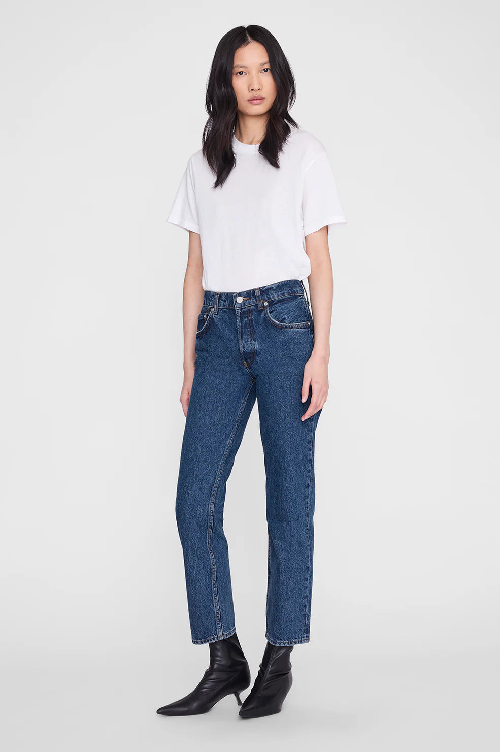 A model wearing a white short-sleeve t-shirt, blue high-waisted straight-leg jeans, and black pointed-toe ankle boots, standing against a light gray studio background.