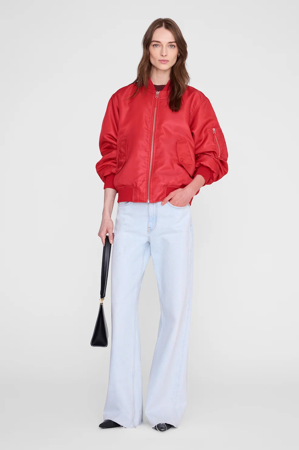 A woman modeling a red satin bomber jacket with light-wash wide-leg jeans and a black handbag, standing against a plain light gray studio background.