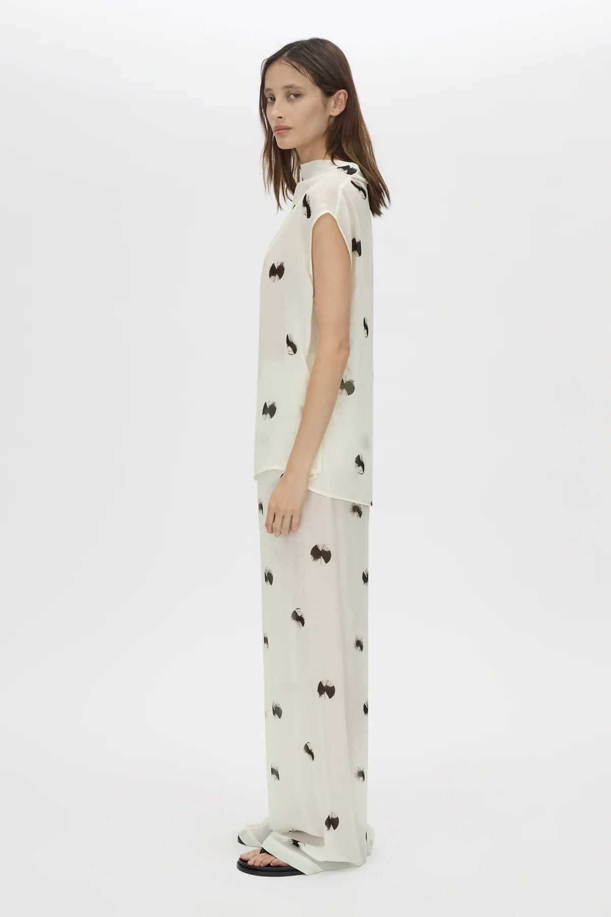 A woman modeling a sleeveless cream-colored dress with a black bow print, shown from a side profile against a plain white studio background.