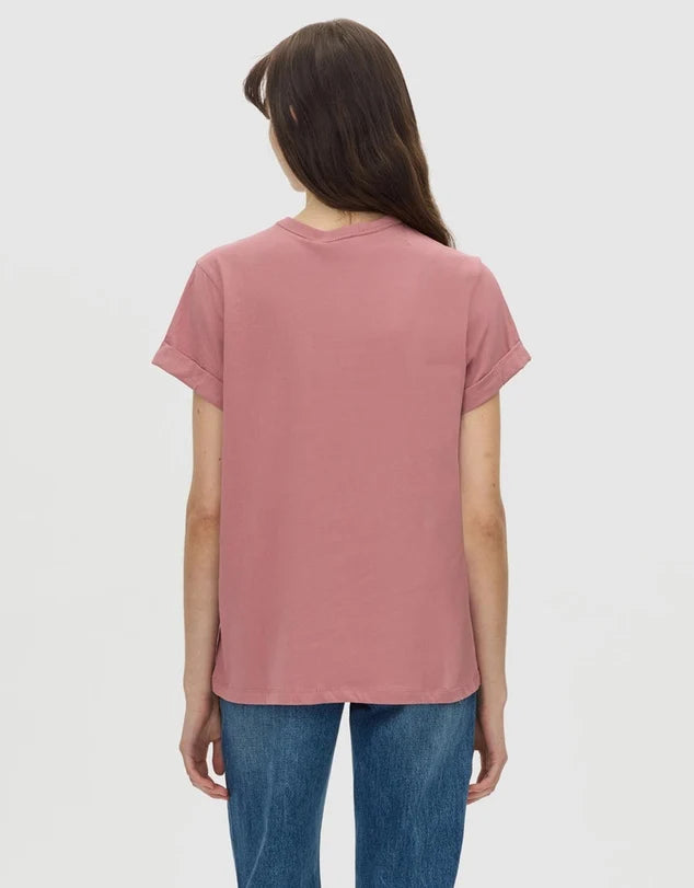 A woman viewed from the back wearing a dusty rose short-sleeve t-shirt and blue jeans against a plain light gray background.