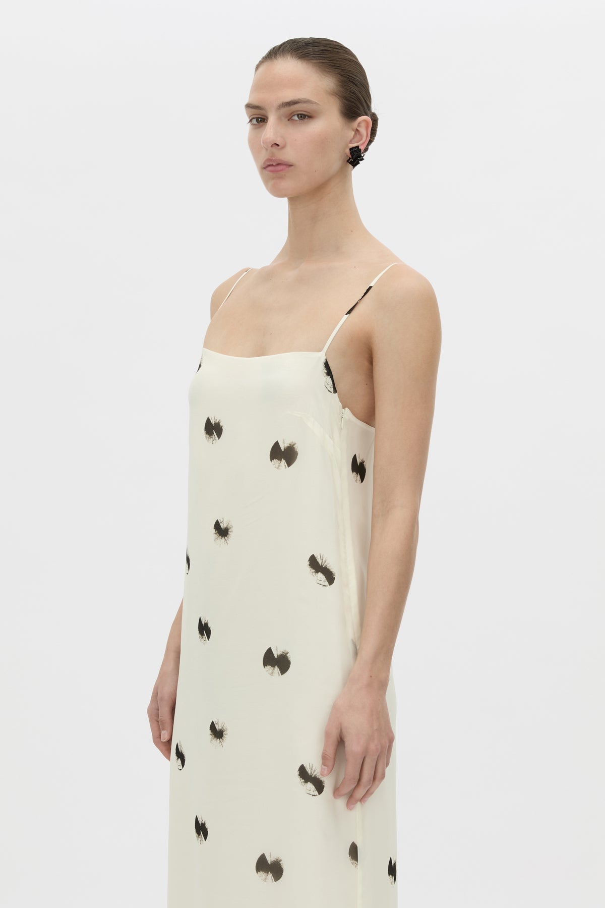 A model wearing a cream-colored spaghetti-strap dress with black bow patterns, photographed against a solid light gray background.