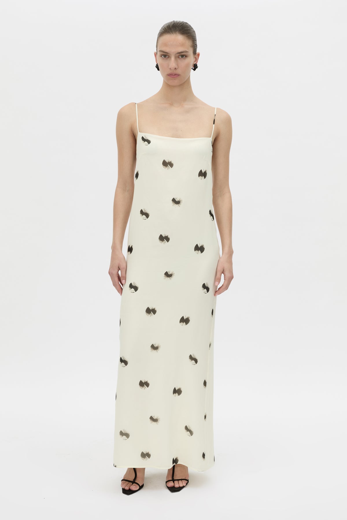 A model wearing a cream-colored maxi dress with thin spaghetti straps and a scattered black bow pattern, paired with black strappy heels, against a plain white studio background.