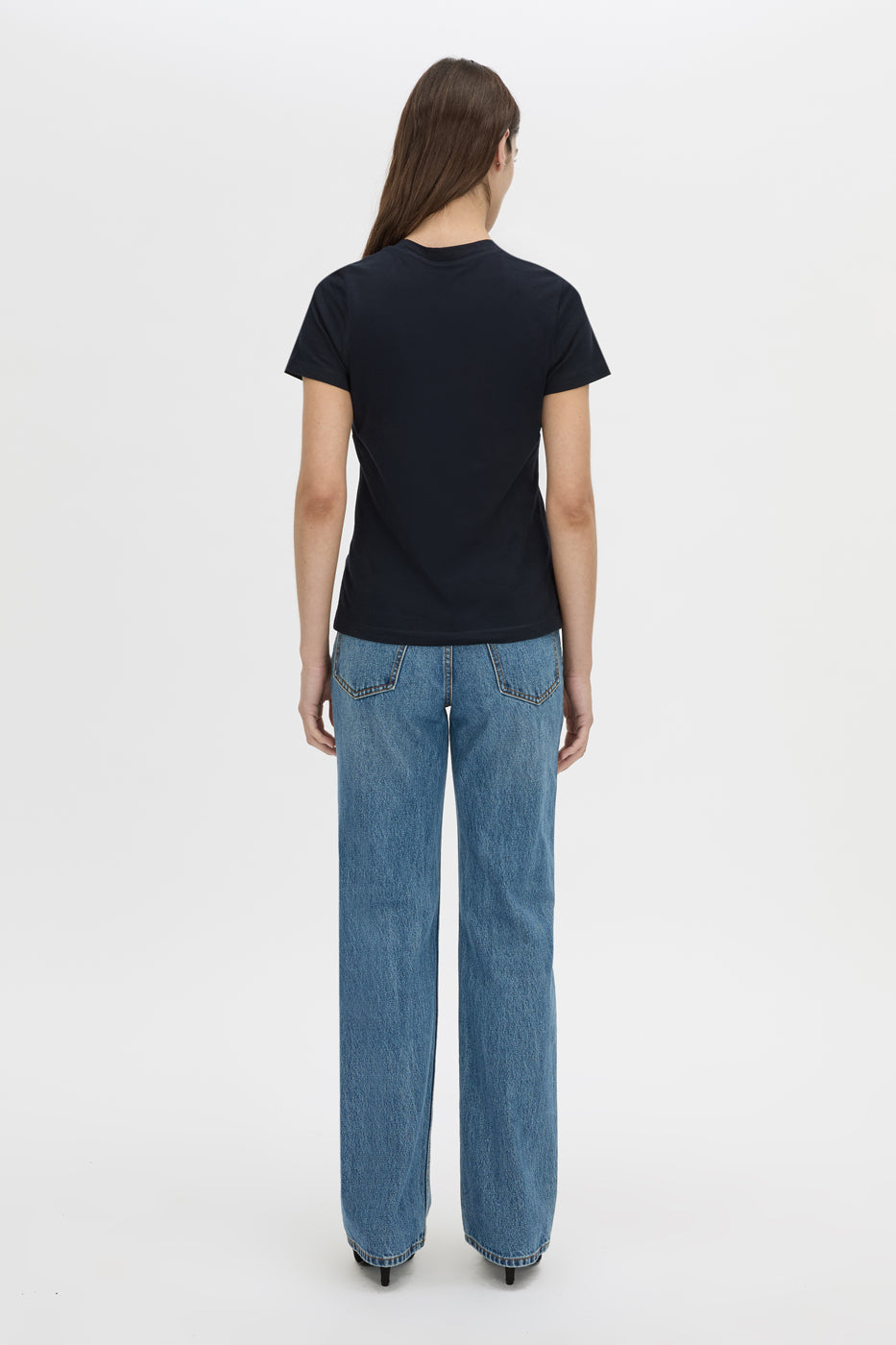 A woman viewed from the back wearing a plain black short-sleeve t-shirt and wide-leg blue jeans, standing against a clean white studio background.