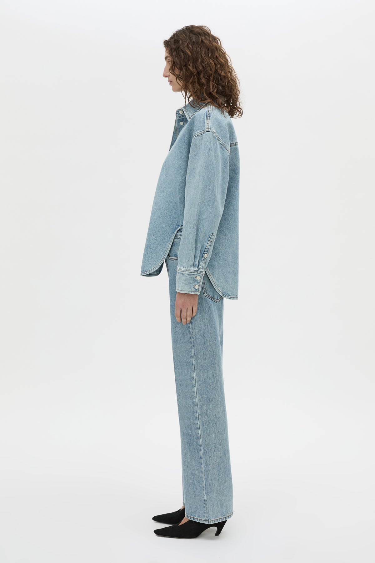A model shown in profile wearing a light blue denim jacket and matching wide-leg jeans, paired with black pointed-toe heels, against a plain white studio background.