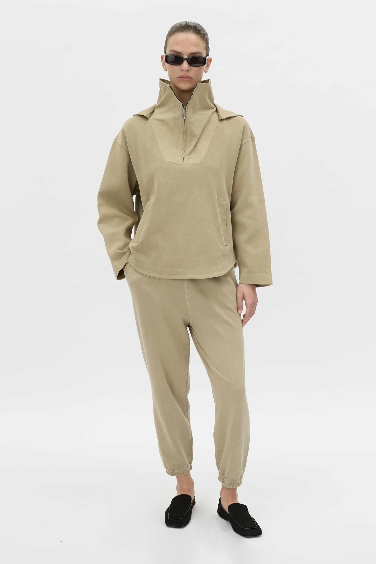A model wearing a matching beige two-piece tracksuit consisting of a high-collar half-zip pullover and jogger pants, styled with black slip-on shoes against a plain white studio background.