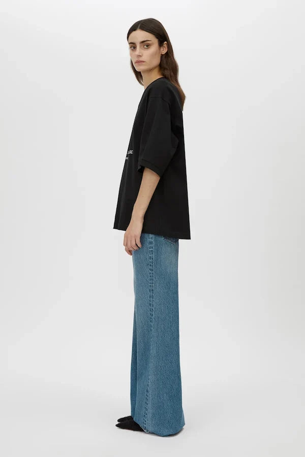 A model is shown from the side wearing a black oversized short-sleeve top and wide-leg blue jeans against a plain light gray studio background.