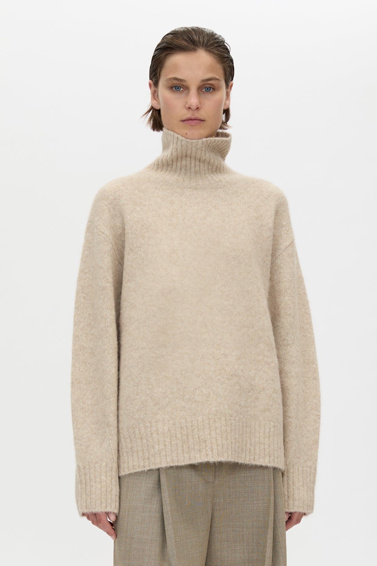 Person wearing a beige turtleneck sweater and pants on a white background