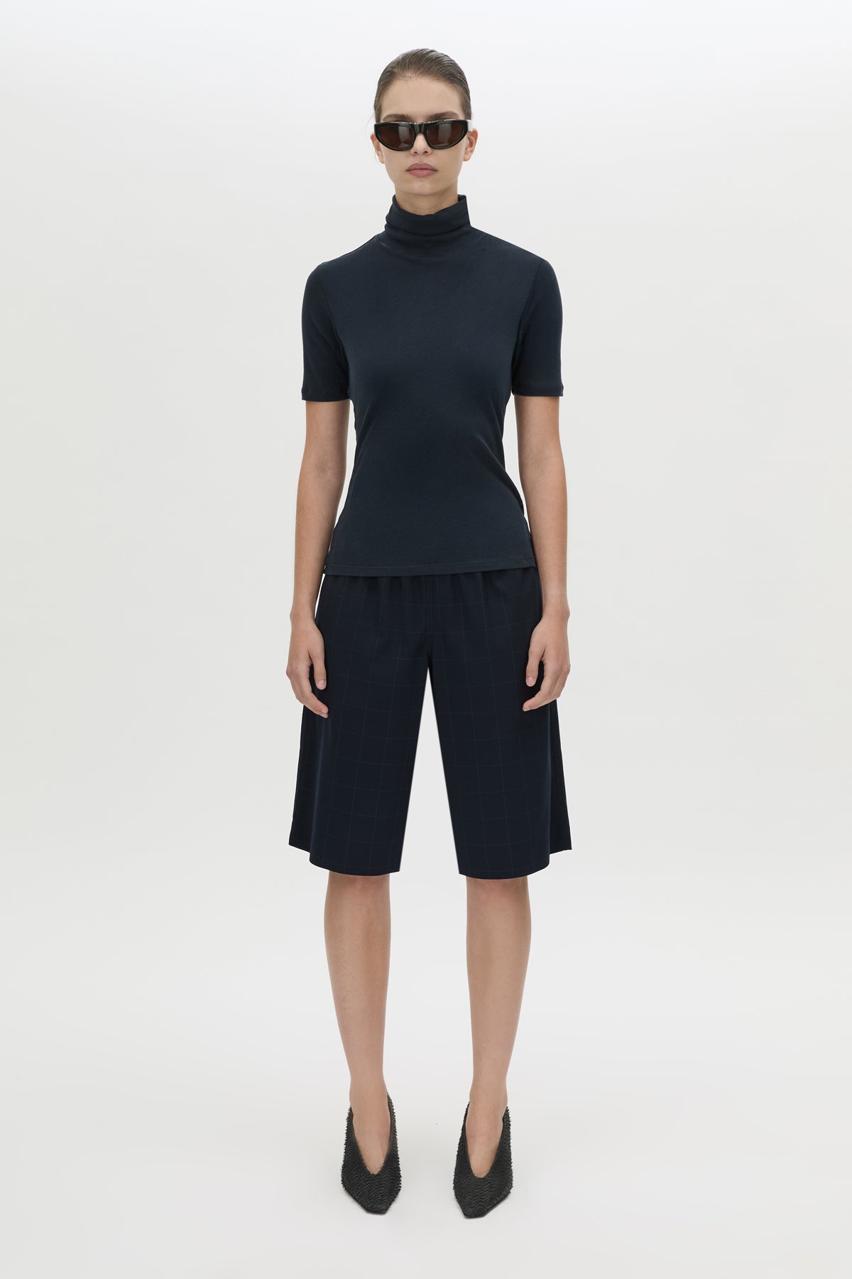 A model stands against a light gray studio background wearing a dark navy short-sleeve mock-neck top and matching dark navy knee-length shorts, with black pointed shoes and sunglasses.