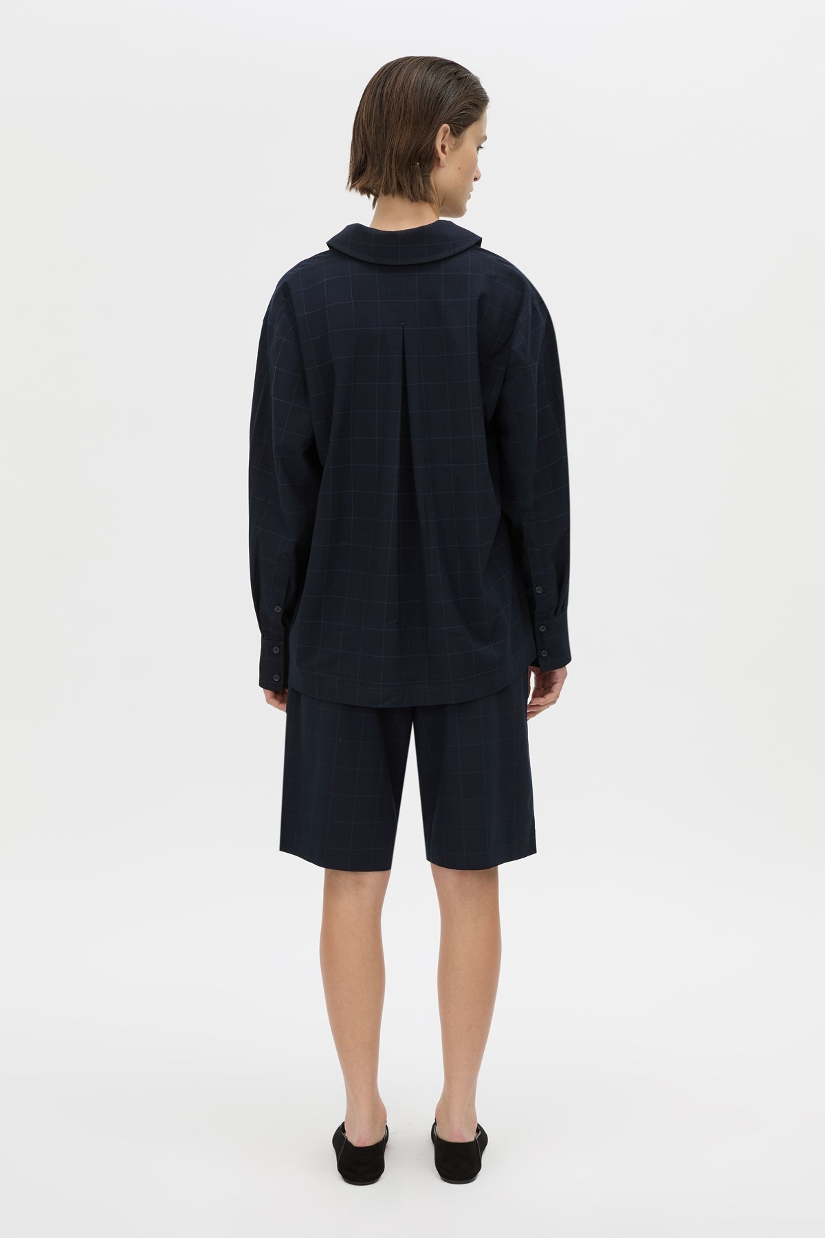 A model viewed from the back wearing a dark navy blue long-sleeve collared shirt and matching shorts, paired with black shoes, against a plain white studio background.