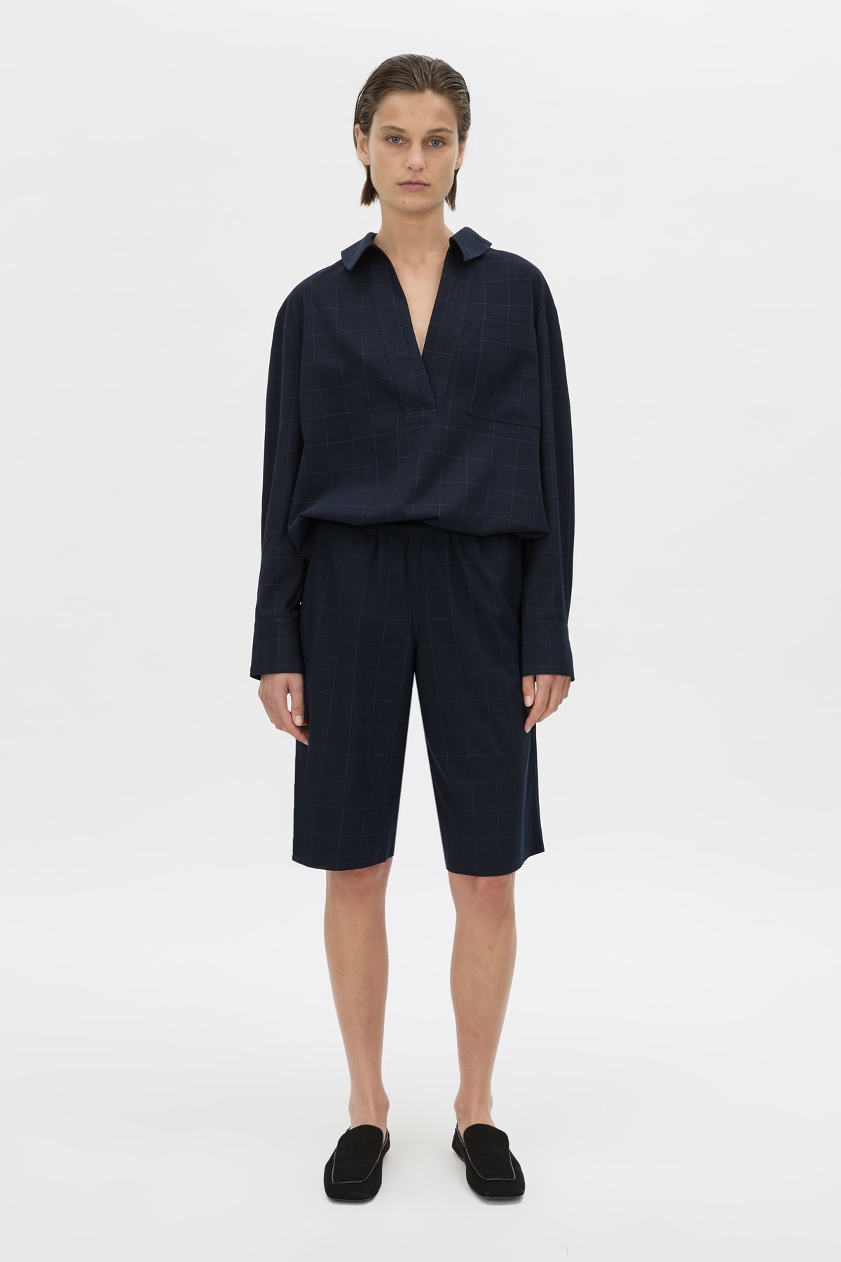 A model stands against a light gray studio background wearing a dark navy long-sleeve collared top and matching dark navy shorts, paired with black slip-on shoes.