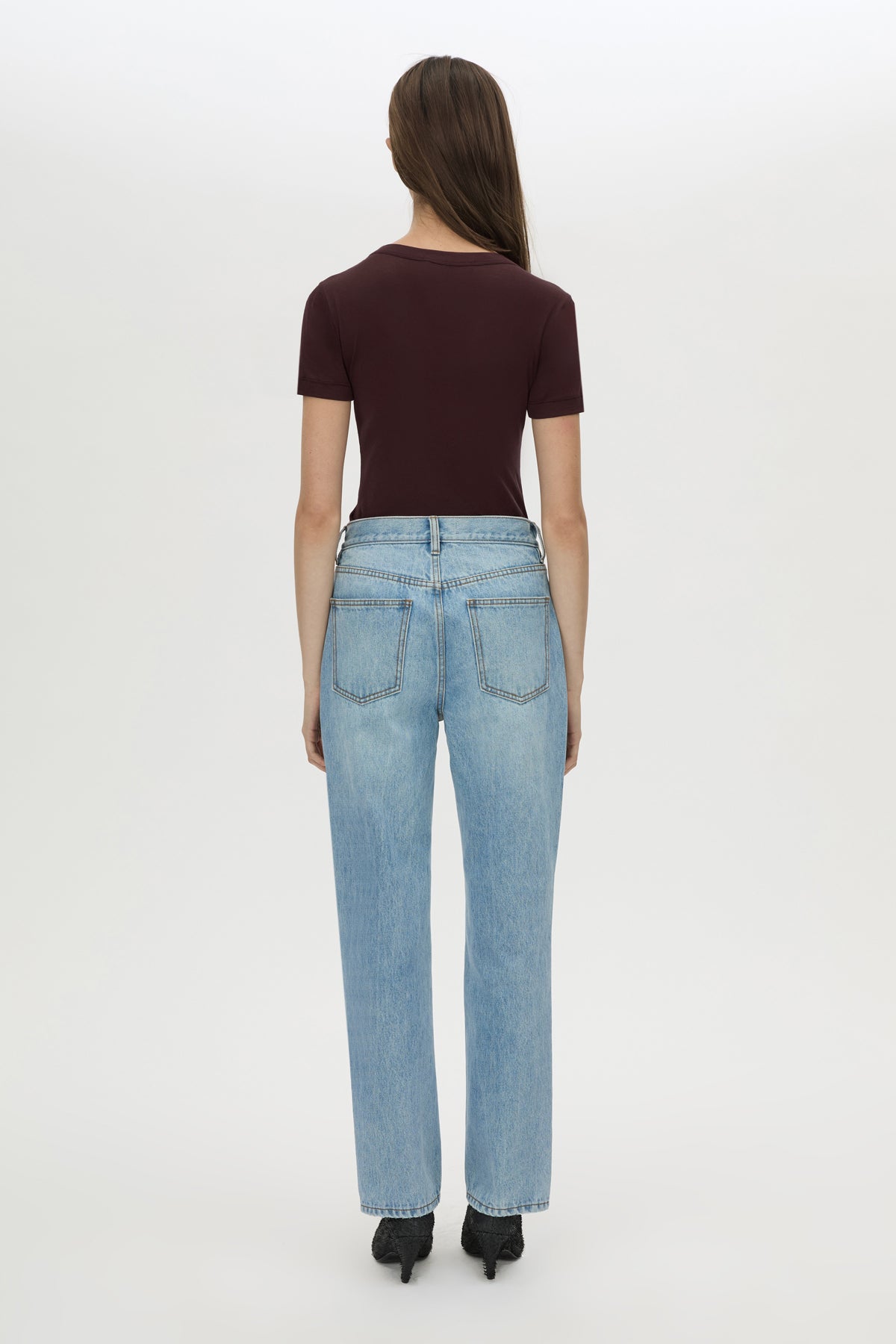 A woman shown from the back wearing a dark burgundy short-sleeve top, light-wash blue jeans, and black heels against a plain white studio background.