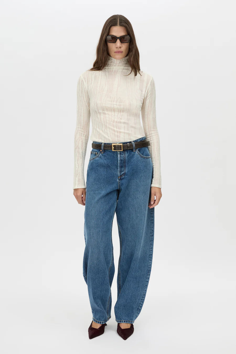 Woman wearing a cream turtleneck, blue jeans, and sunglasses on a white background