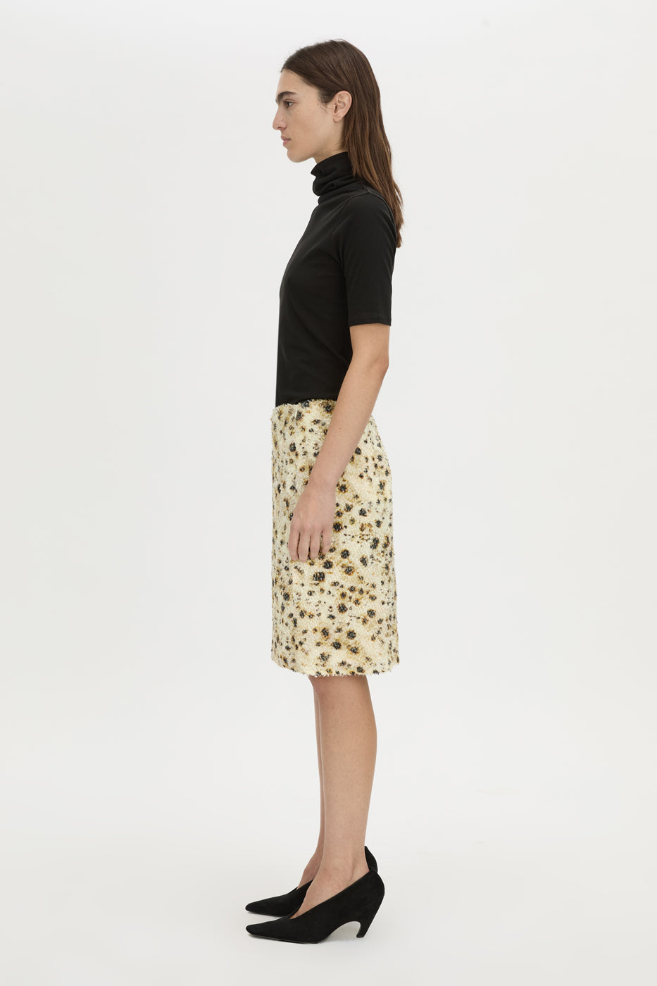 A woman shown in profile wearing a black short-sleeve mock-neck top and a knee-length cream skirt with a small floral print, paired with black pointed-toe heels, against a plain white studio background.