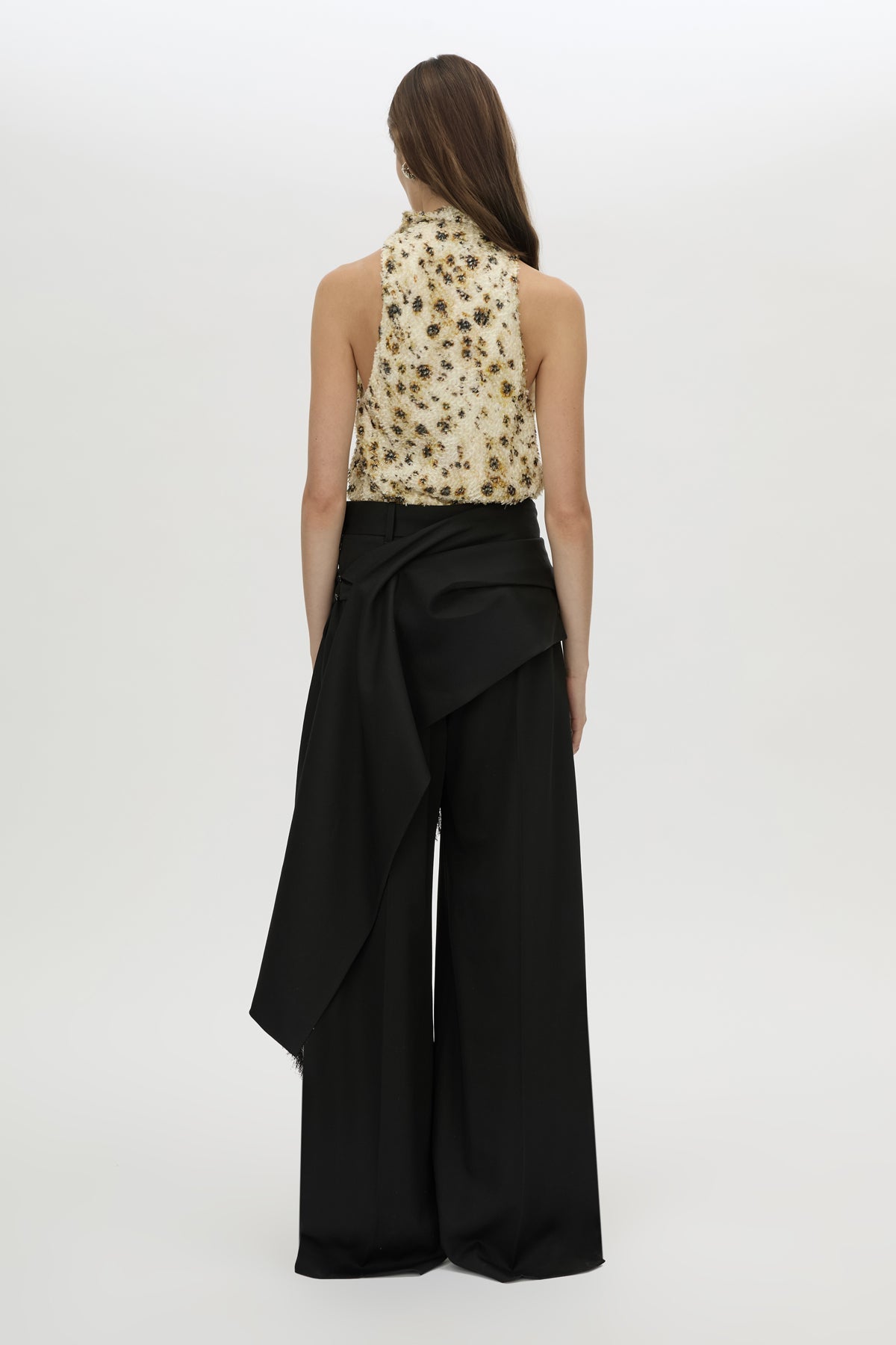 A model shown from the back wearing a sleeveless beige top with a black floral pattern and high-waisted black wide-leg draped trousers, set against a plain light gray studio background.