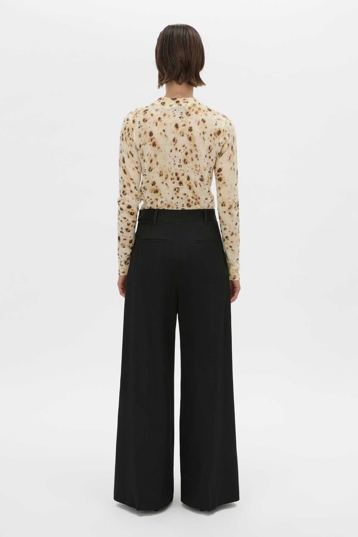 A model viewed from the back wearing a cream-colored long-sleeve blouse with a brown floral pattern and black wide-leg trousers, set against a plain white studio background.