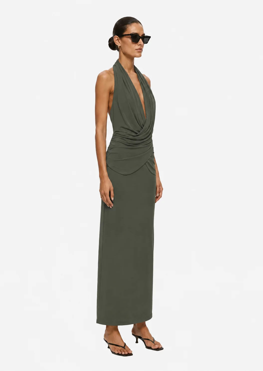 A woman modeling a sleeveless olive green halter maxi dress with a draped wrap-style bodice and deep V-neckline, paired with black strappy heels and sunglasses, against a plain light gray studio background.