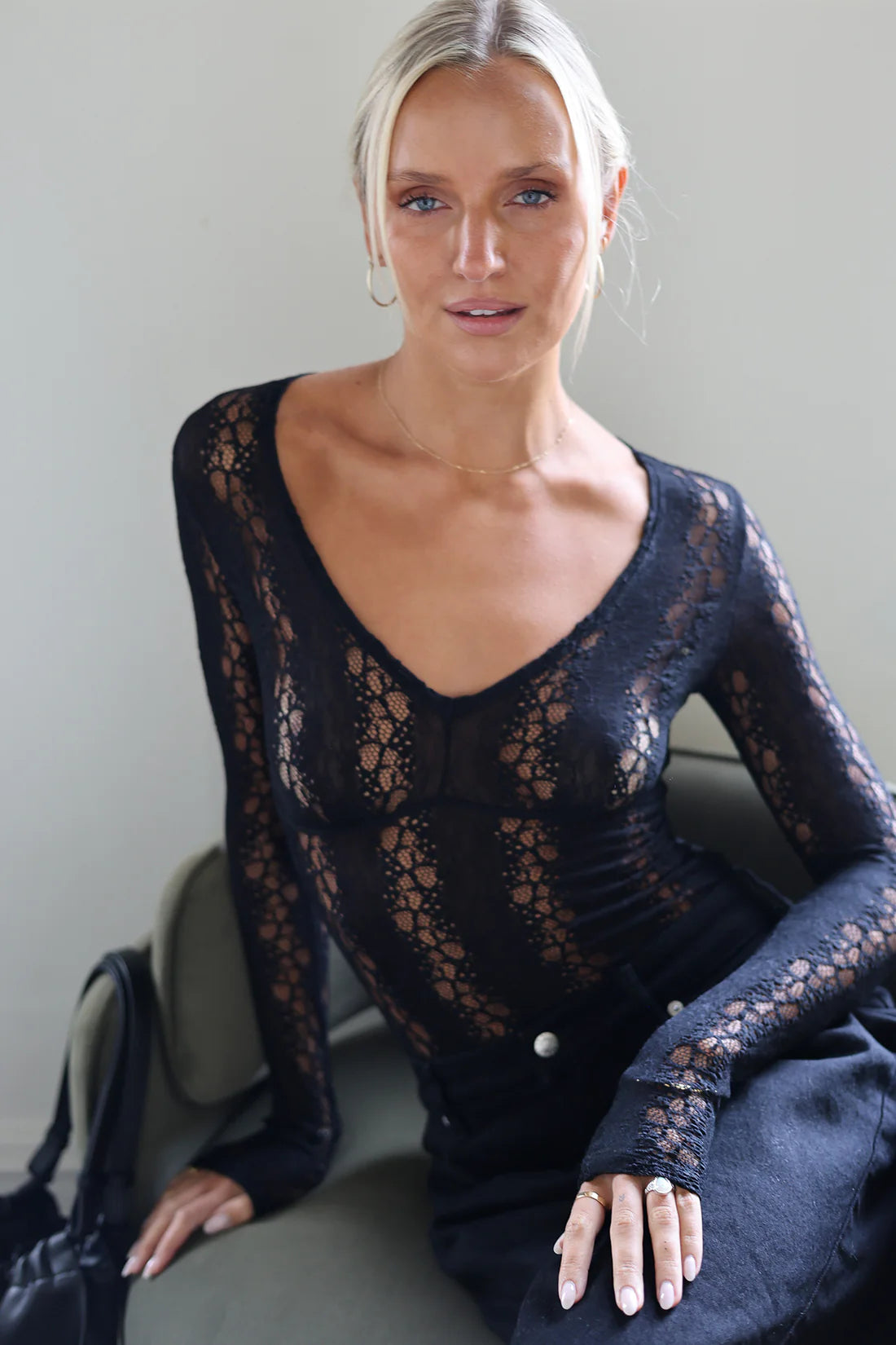A blonde woman wearing a black lace long-sleeve bodysuit with a deep V-neck, seated on a gray chair against a neutral gray background.