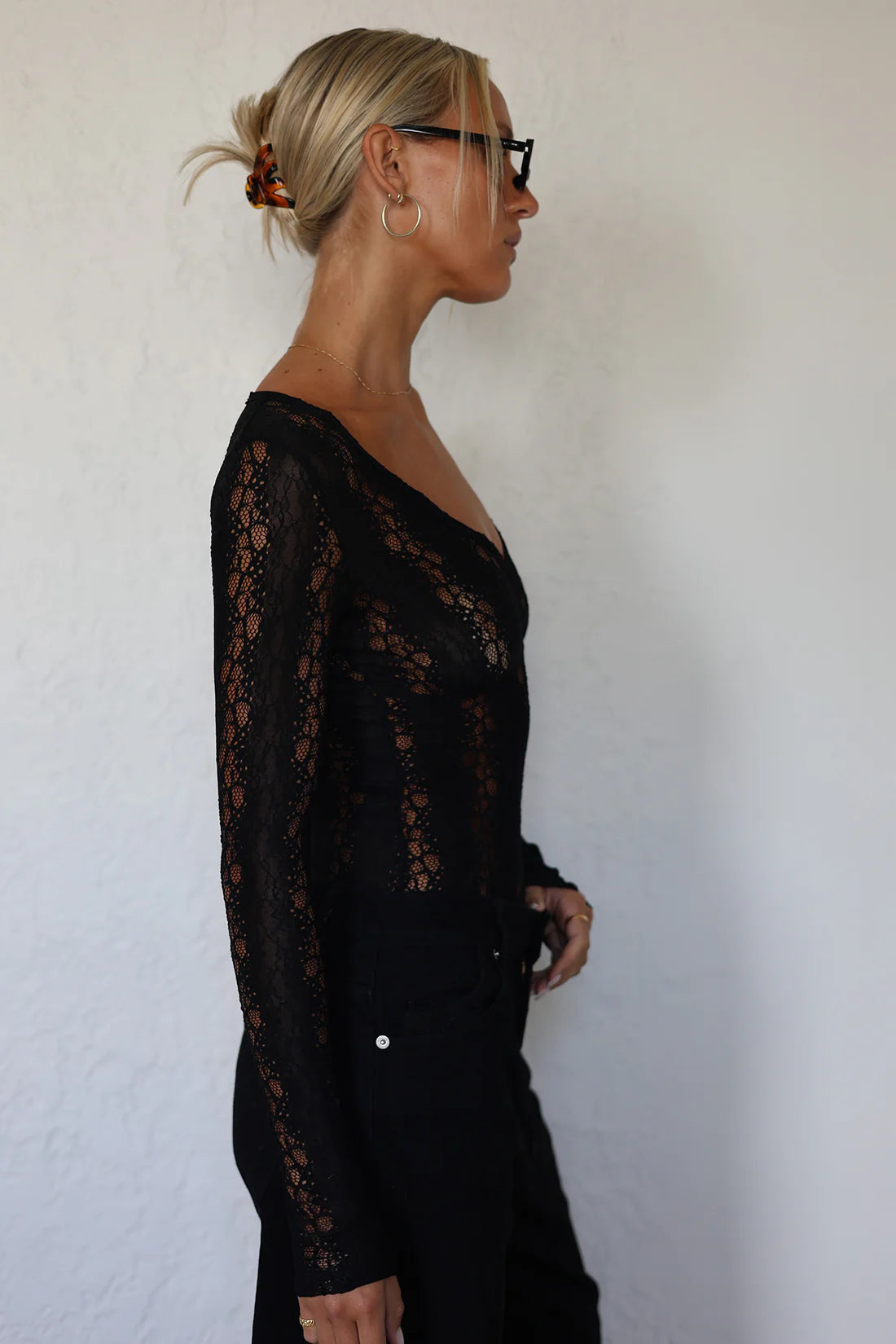 A woman in profile wearing a black lace long-sleeve top and black pants, standing against a plain off-white wall.