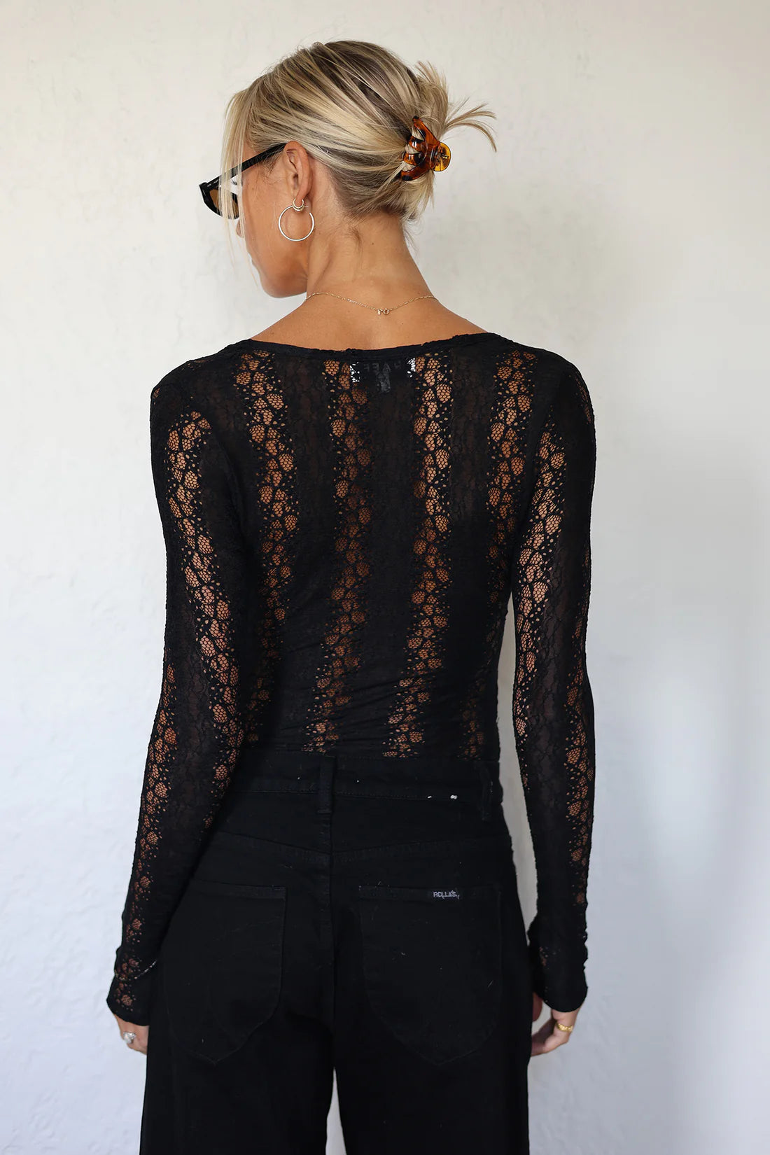 A woman shown from the back wearing a black sheer lace long-sleeve top and black pants against a plain light wall.