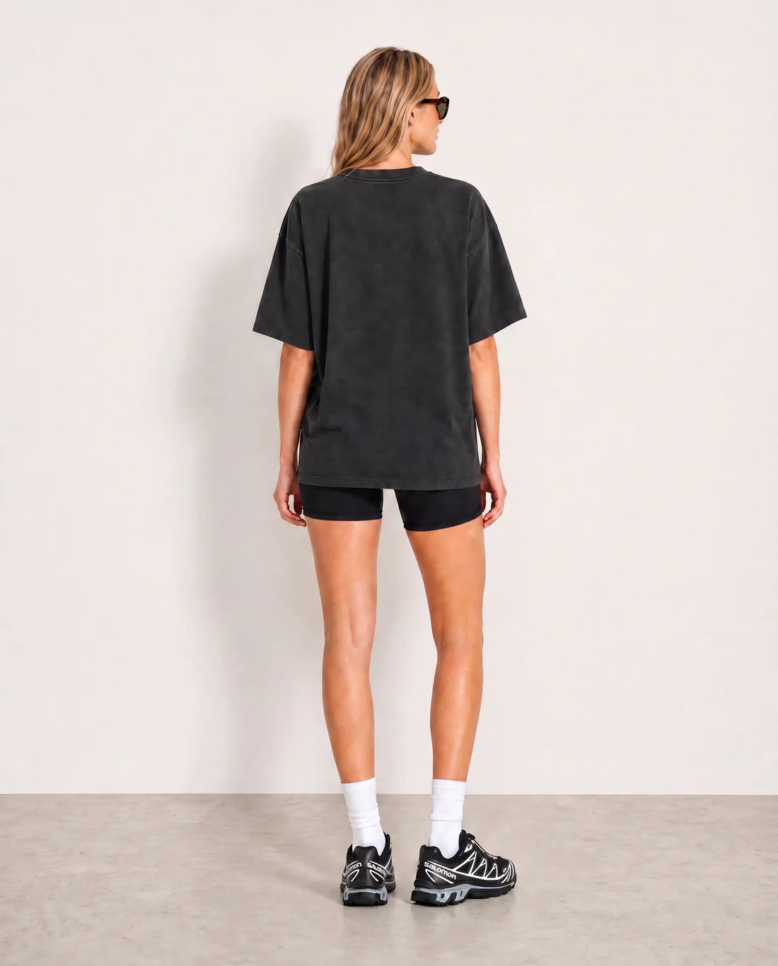 A woman is shown from behind wearing an oversized black washed t-shirt, black bike shorts, white socks, and black and white sneakers against a minimalist off-white studio background.