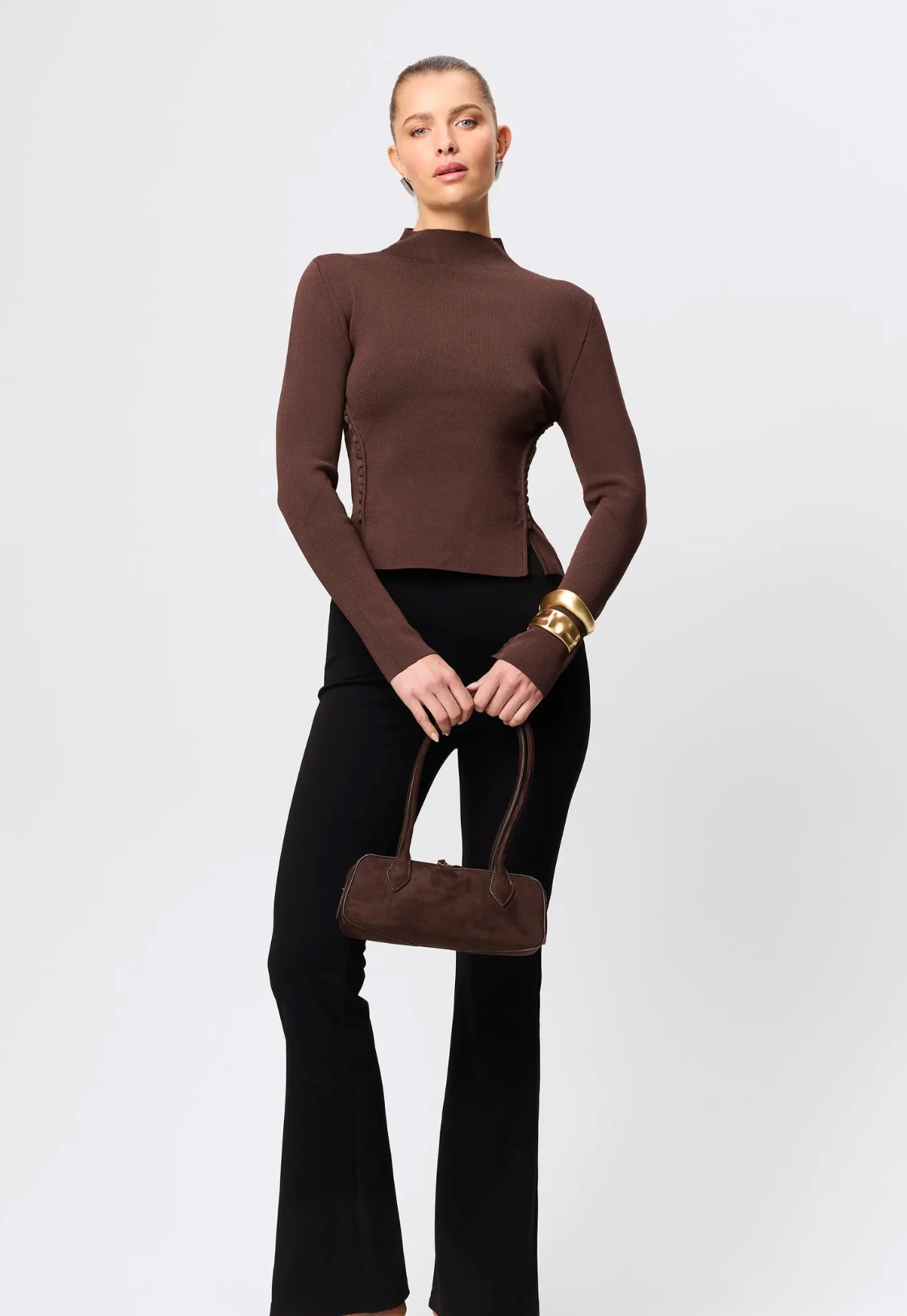 Woman wearing a brown long-sleeve top and black pants, holding a brown handbag on a white background