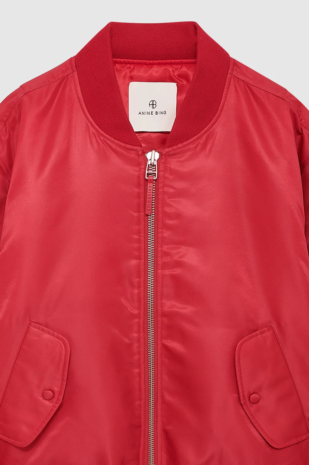 A close-up of a red nylon bomber jacket with a silver zipper, ribbed collar, and front flap pockets, showing the Anne B65 brand label inside the neck.