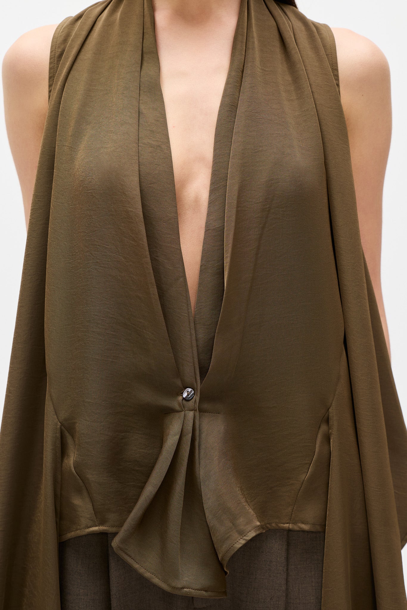 A close-up of a model wearing a sleeveless, deep V-neck draped top in olive brown. The top features a single button closure near the center and is styled over dark trousers against a plain light background.