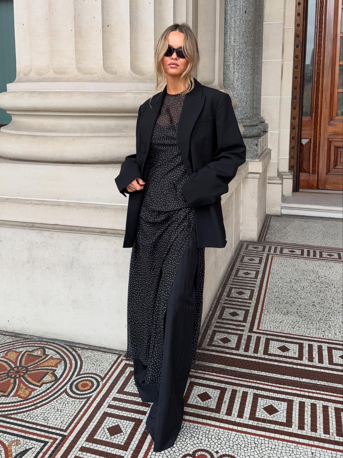 A woman stands outdoors in front of a stone building, wearing a black oversized blazer over a black patterned maxi dress and black wide-leg trousers, with dark sunglasses.