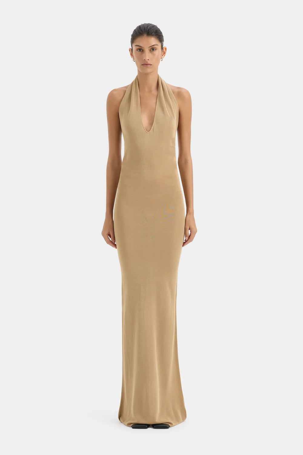 A model wearing a sleeveless, halter-neck, floor-length beige dress against a plain light gray studio background.