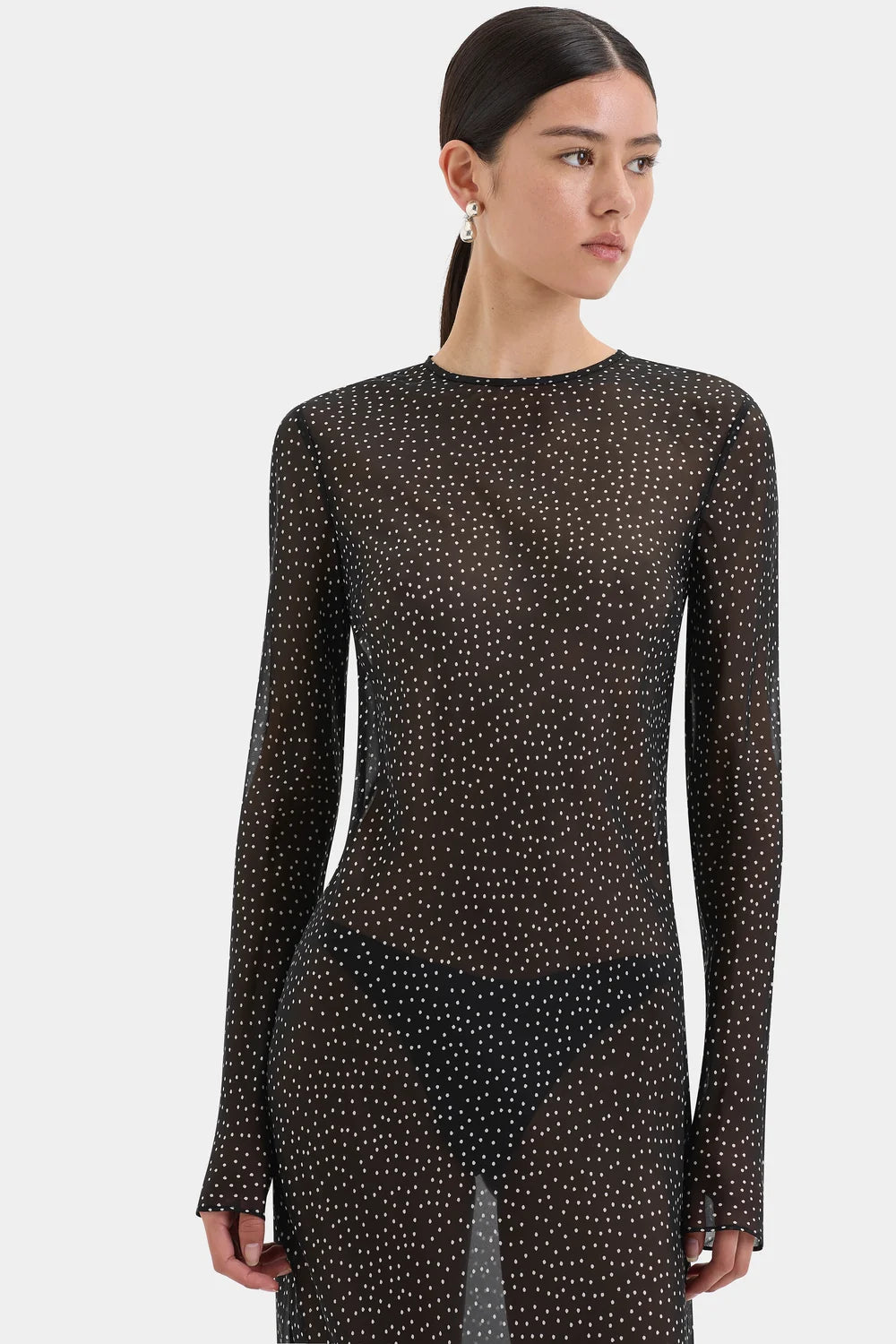 A model wearing a sheer black long-sleeve top and matching skirt with a small white polka dot pattern, photographed against a light gray studio background.