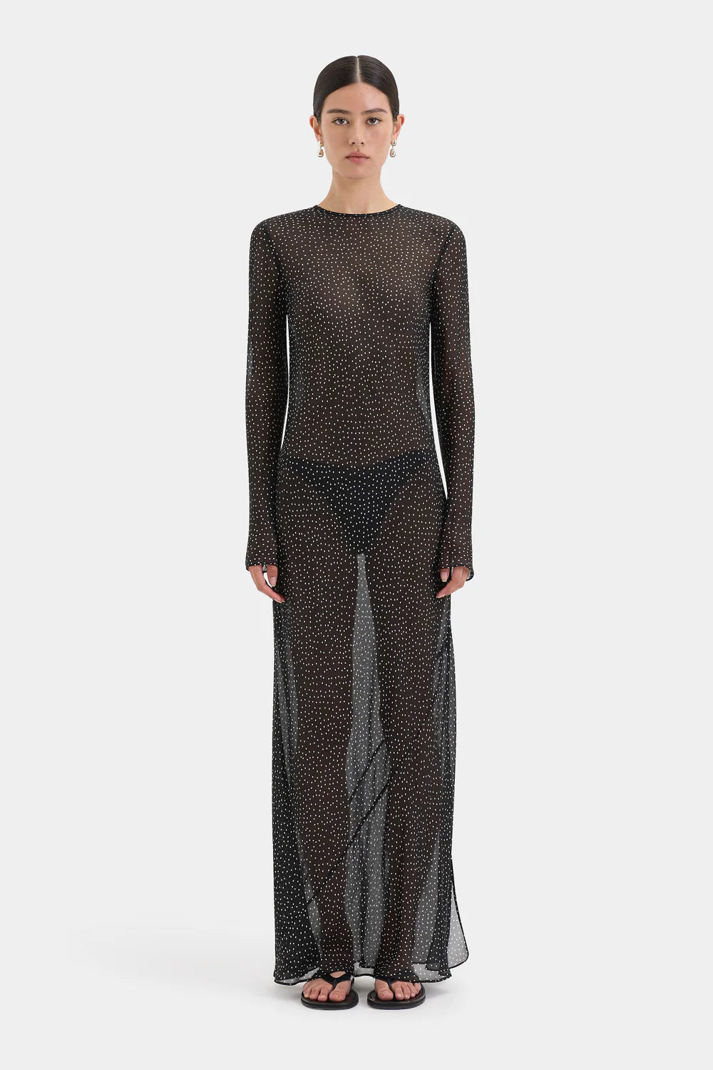 A model wearing a sheer, long-sleeved maxi dress with a dense black and brown abstract pattern, standing against a light gray studio background.