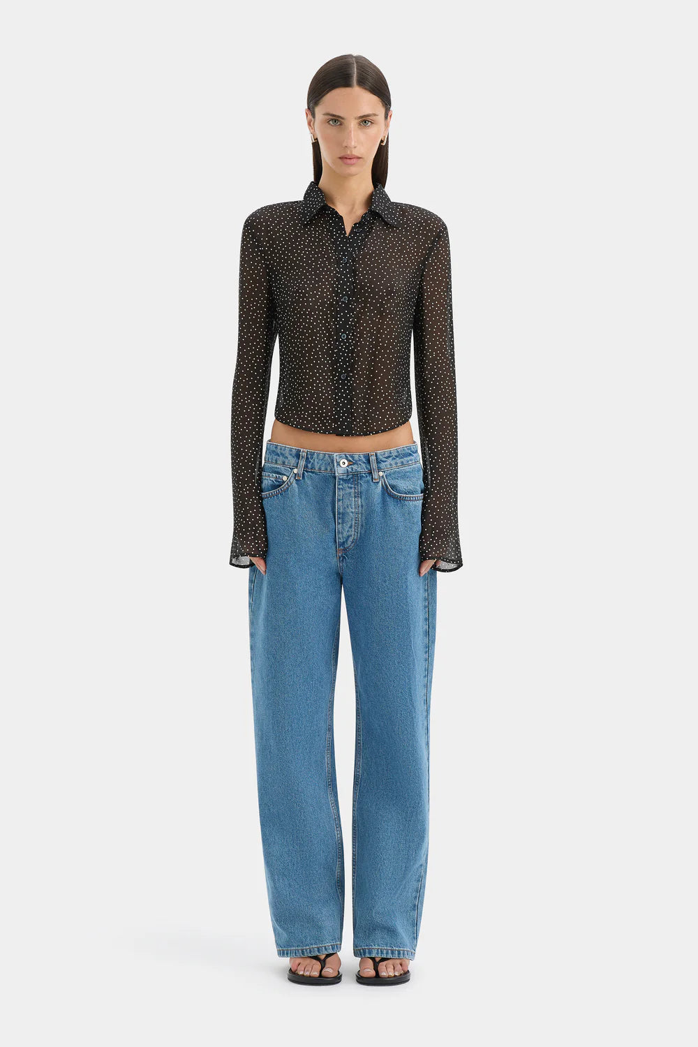 A model stands against a light gray studio background wearing a sheer black long-sleeve button-up shirt with a small repeating pattern, paired with wide-leg blue jeans and black sandals.