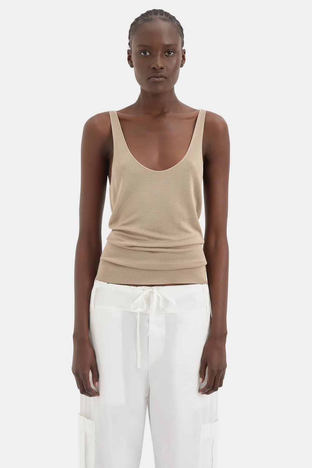 A model wearing a beige ribbed knit scoop-neck tank top and white drawstring pants, standing against a light gray studio background.