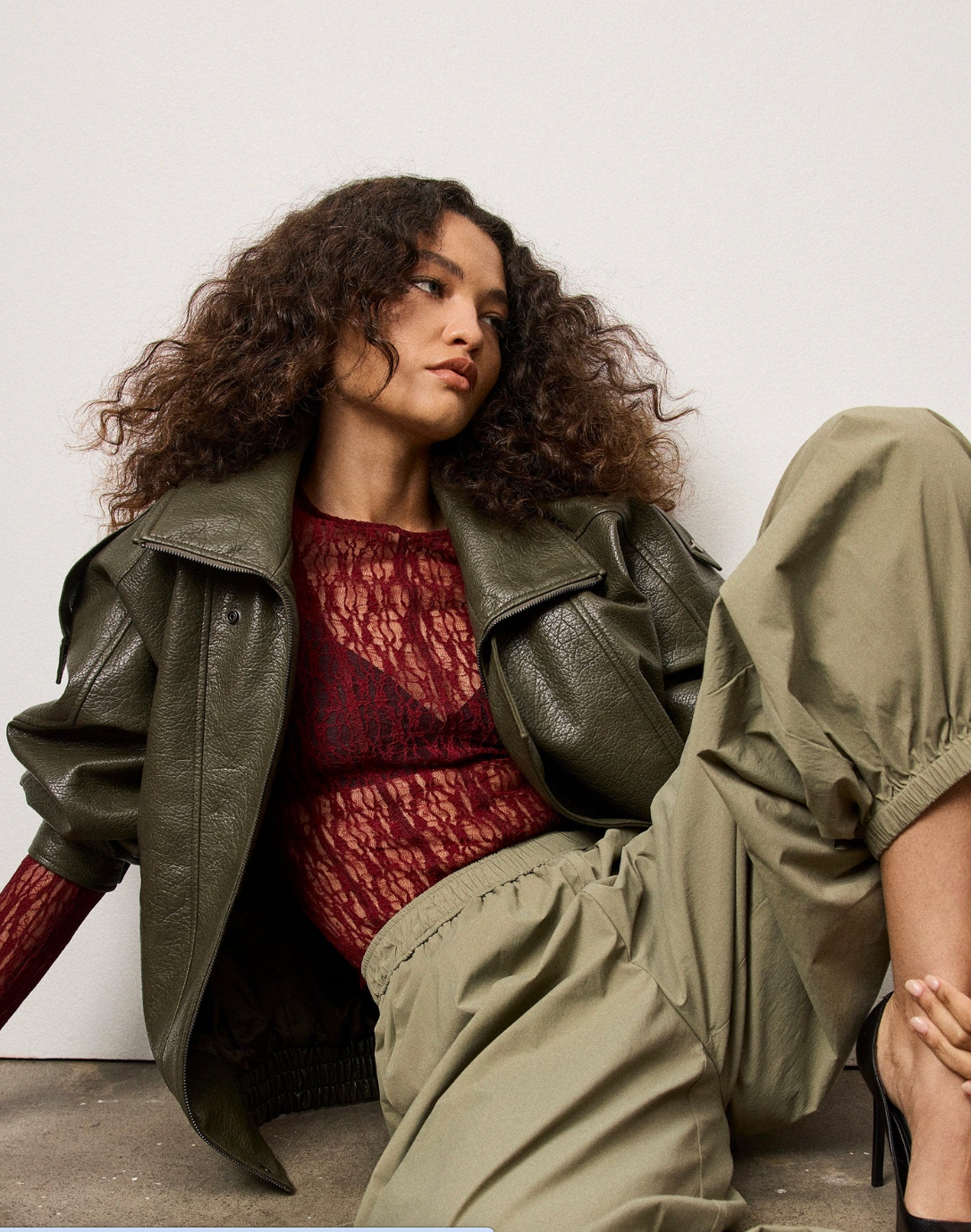 Woman wearing a green jacket and red top sitting on a white floor.
