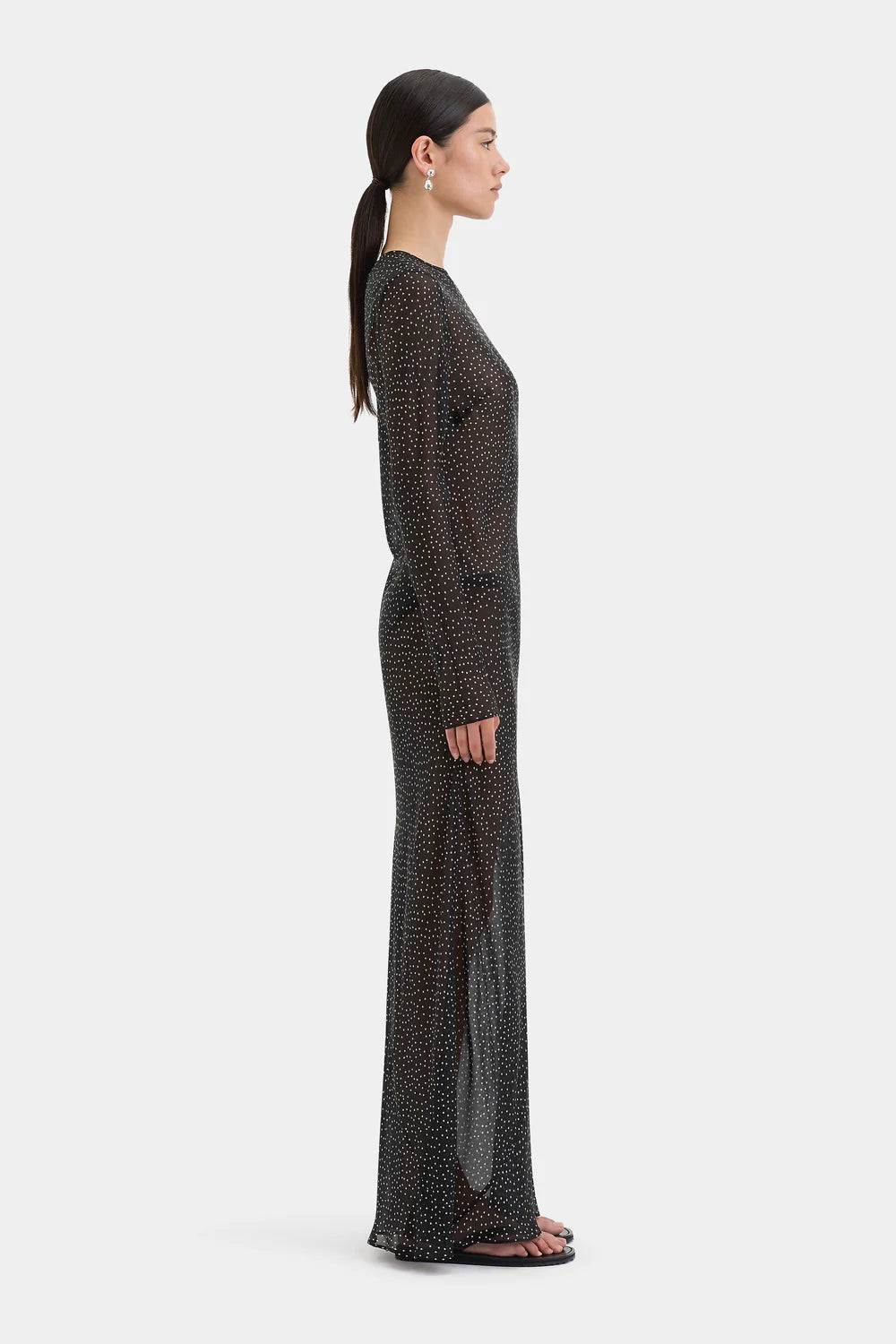 A woman shown in side profile wearing a long-sleeve, floor-length sheer maxi dress with a small black-on-white allover print, set against a solid light gray background.