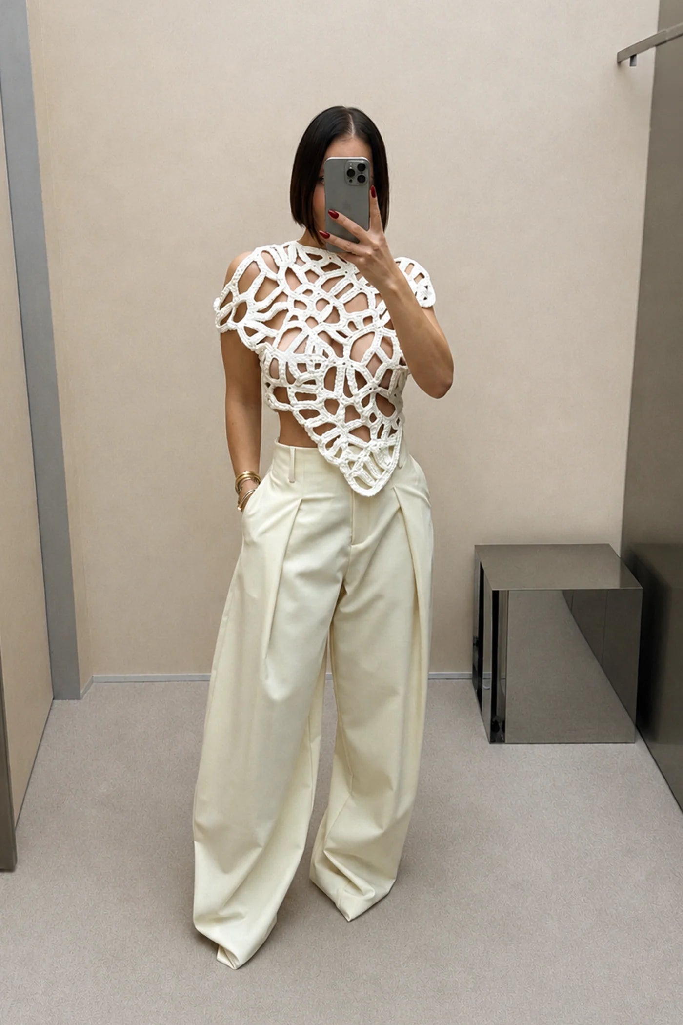 A woman takes a mirror selfie in a fitting room wearing a white short-sleeve crochet cutout crop top and cream wide-leg trousers.