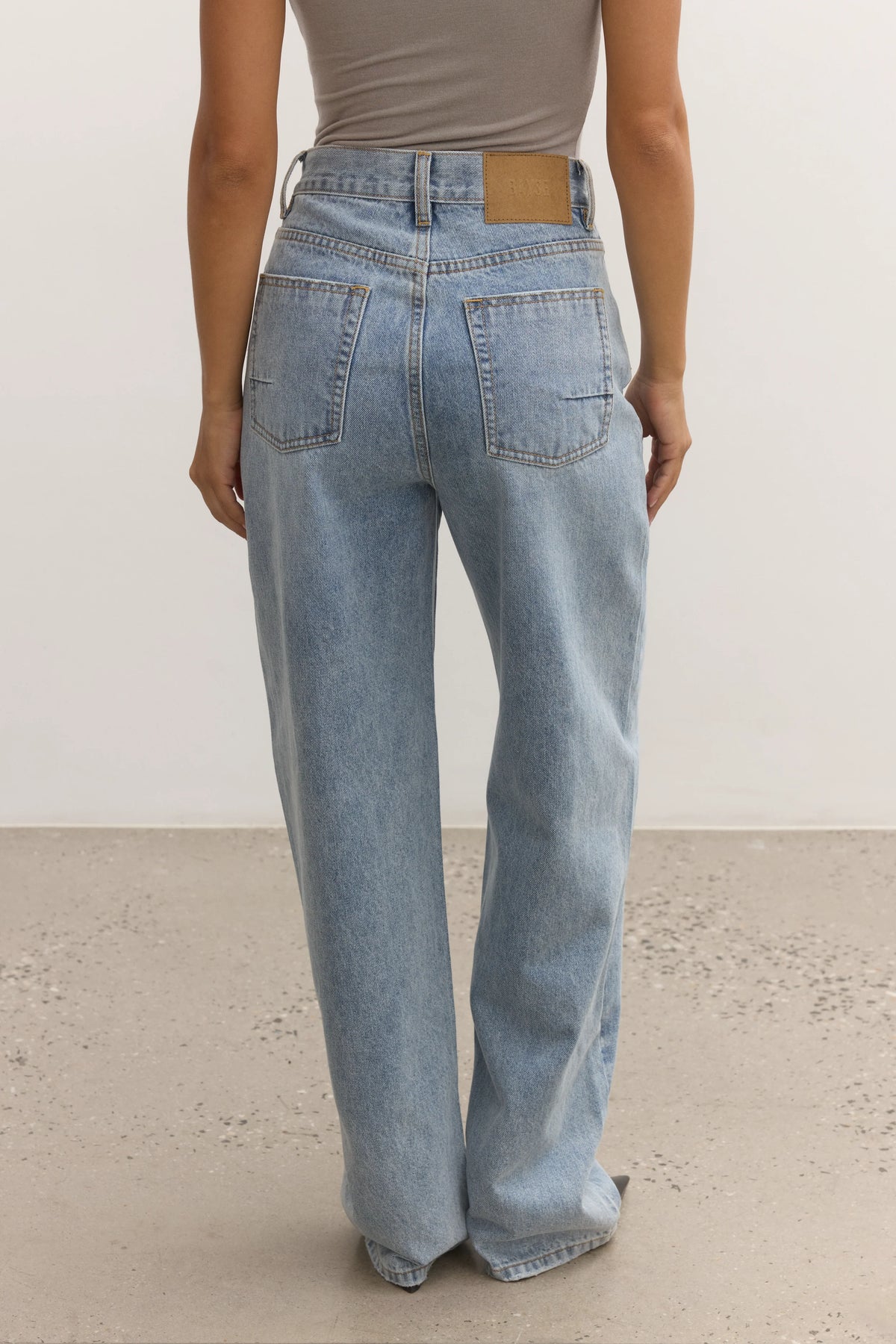 A person shown from the back wearing light-wash wide-leg jeans and a taupe top, standing against a plain off-white wall and light concrete floor.