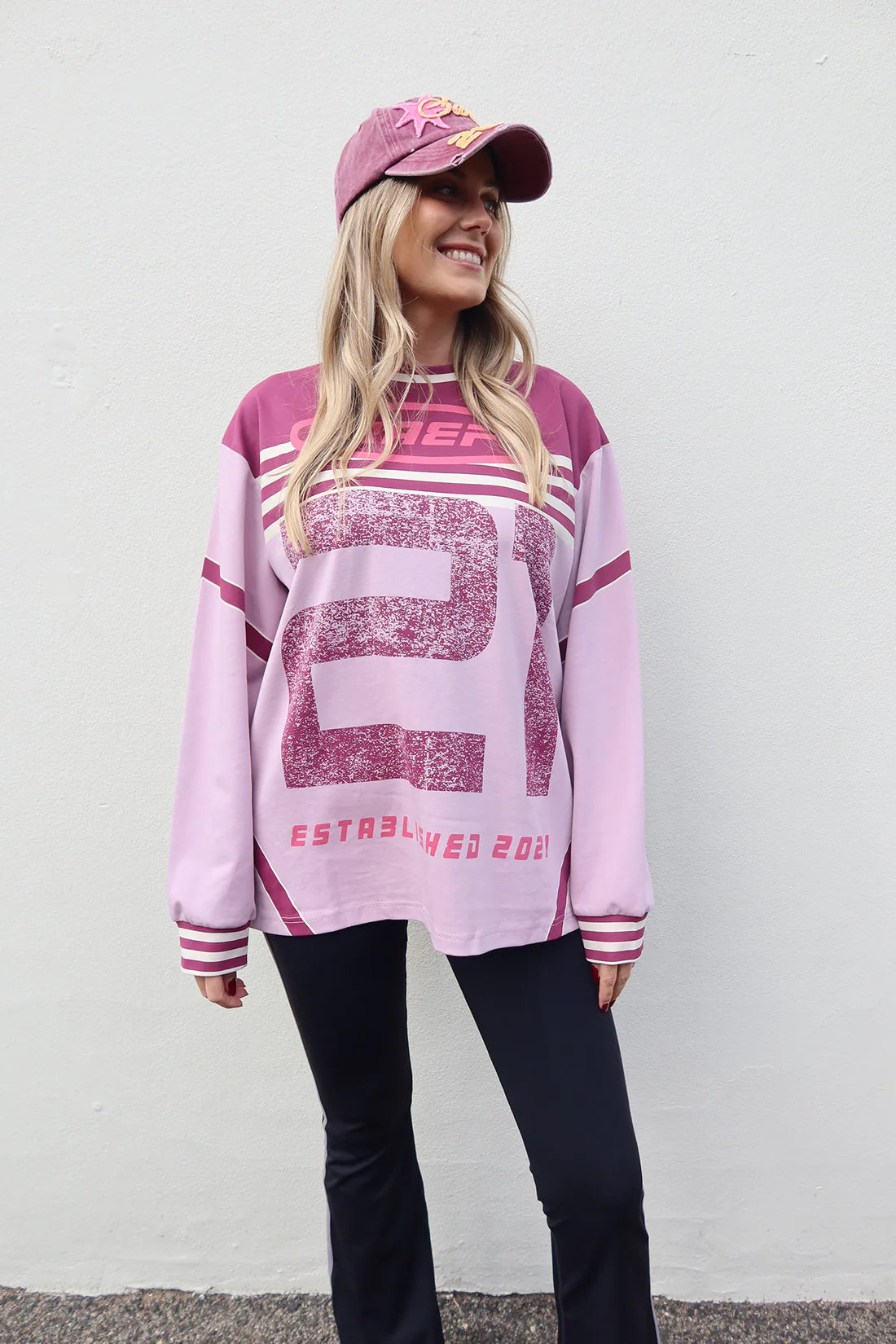 A woman wearing a light pink and dark pink long-sleeve athletic-style sweatshirt with a large distressed number 21 on the front, paired with black pants and a matching pink cap, standing against a plain white wall.