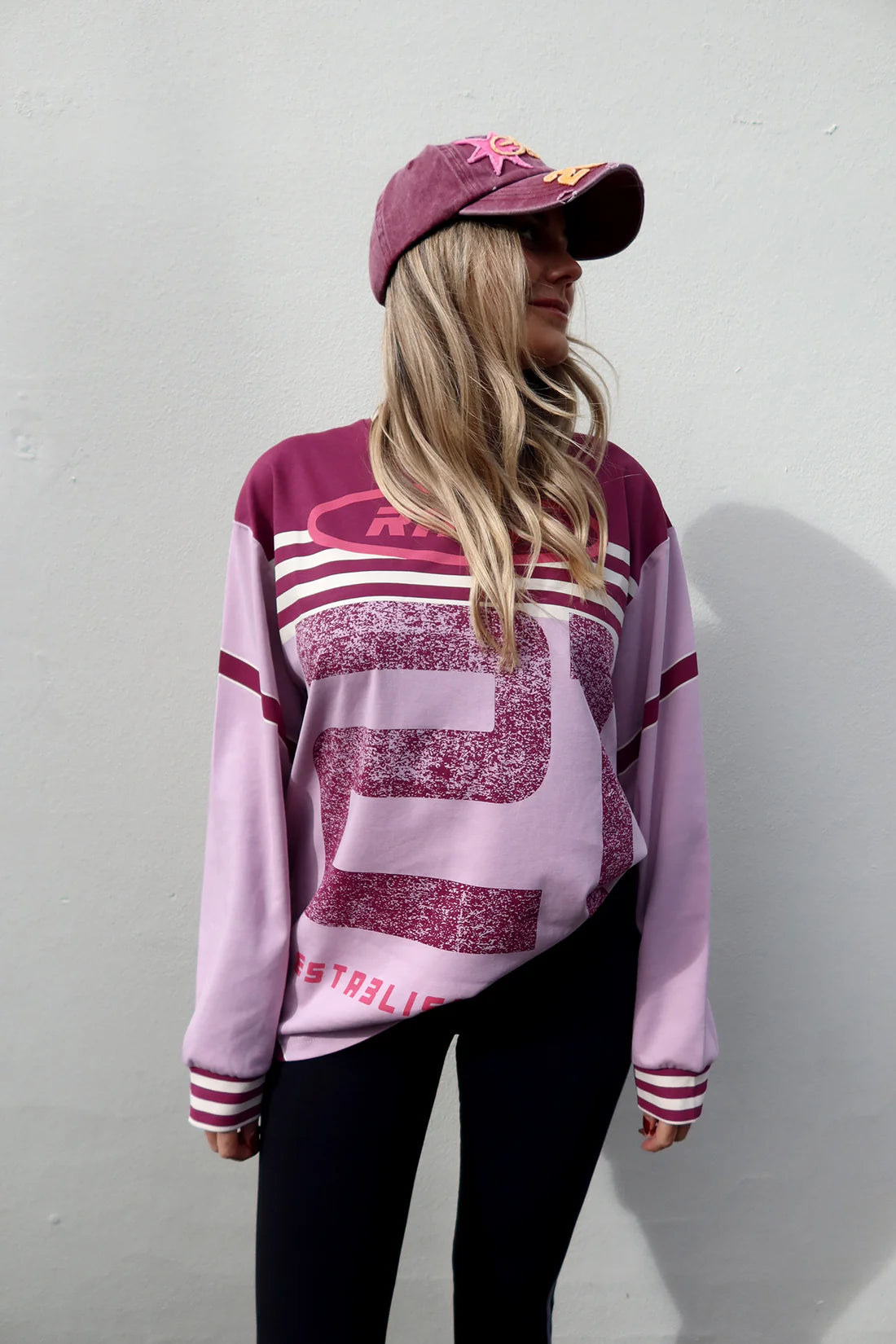 A woman wearing a light pink and maroon long-sleeve athletic-style sweatshirt with a large distressed number 9 graphic, paired with black leggings and a maroon baseball cap, standing against a light gray wall.