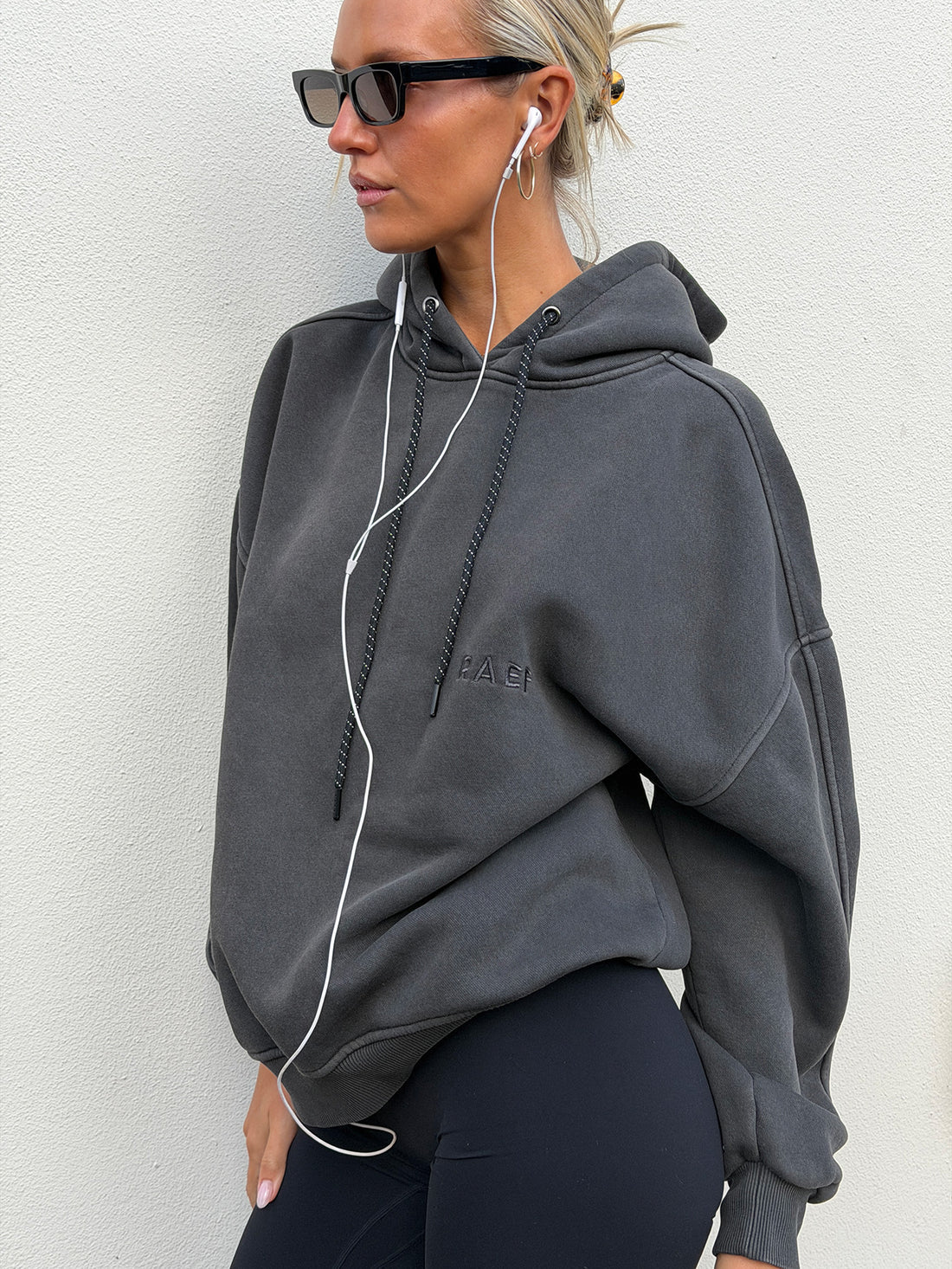 A woman wearing a dark gray oversized hoodie with a tonal embroidered logo on the chest, black sunglasses, and white wired earphones, standing against a light gray textured wall.