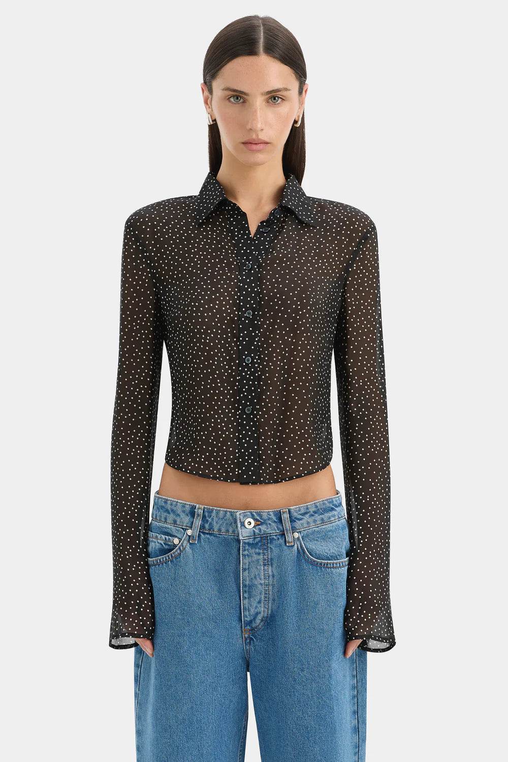 A model wearing a black long-sleeve sheer button-up shirt with a small white polka dot pattern, paired with blue jeans, against a light gray background.