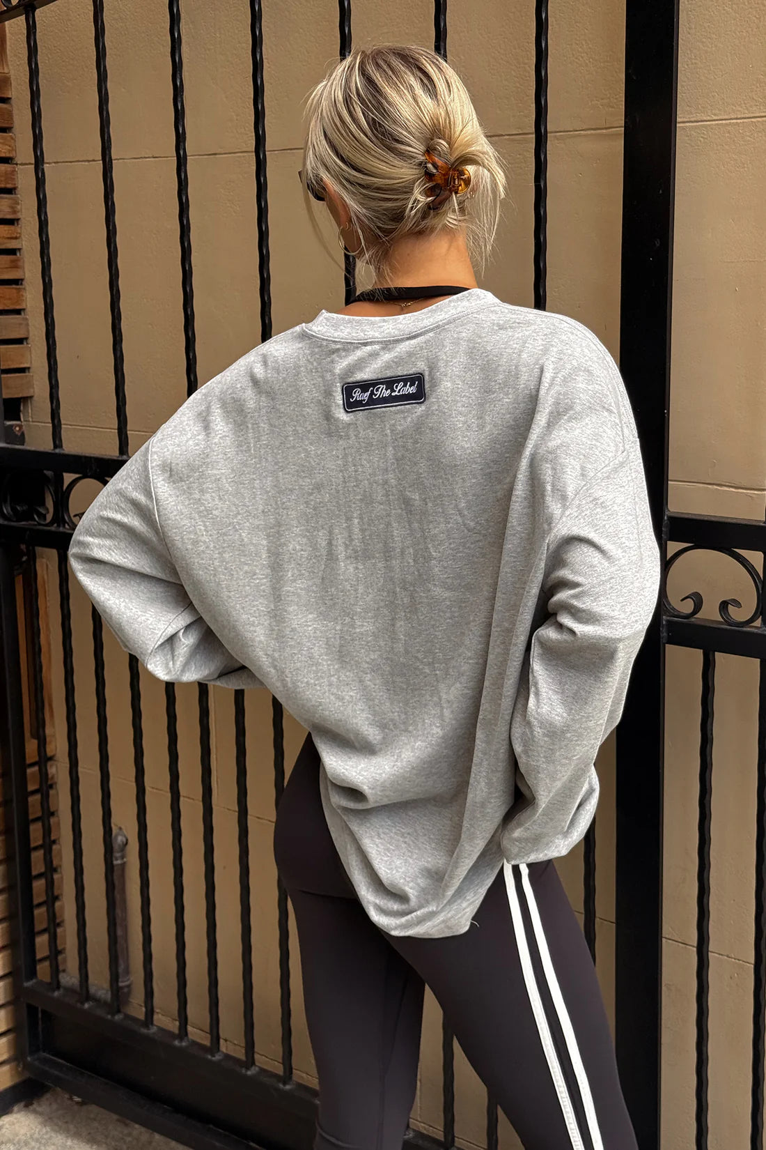 A woman viewed from behind wearing a light gray oversized crewneck sweatshirt with a small black brand patch on the back, paired with black leggings featuring white side stripes, standing in front of a beige wall and black metal gate.
