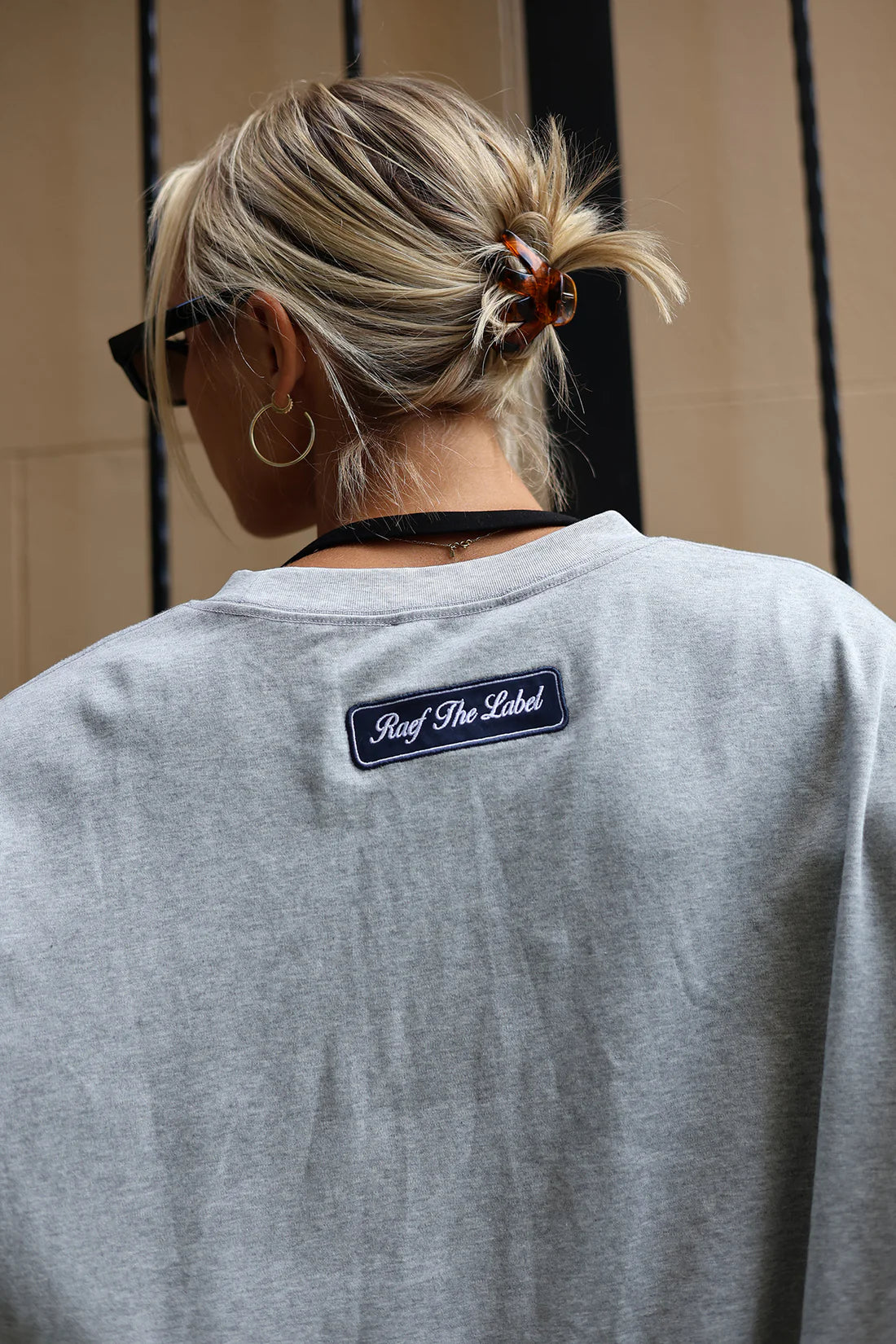 A person viewed from behind wearing a light gray heather crewneck sweatshirt with a 'Surf The Label' patch on the back, set against a neutral indoor background.