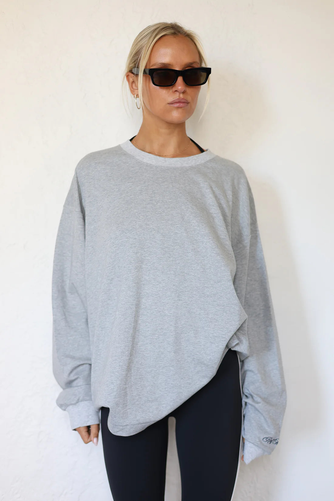 A woman modeling a light gray oversized long-sleeve sweatshirt with an asymmetrical hem, paired with black leggings, against a white textured wall.