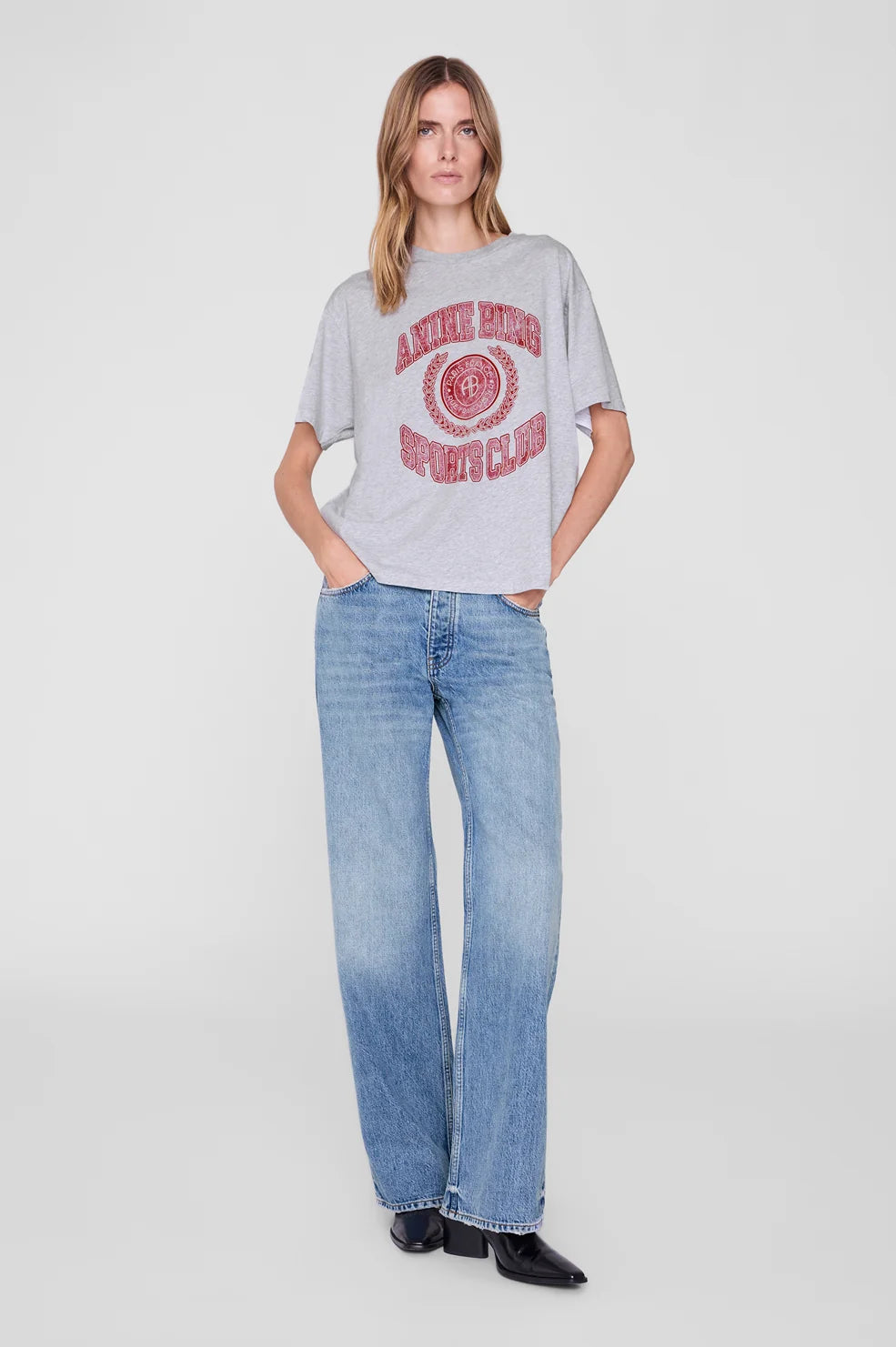 A woman modeling a light gray oversized graphic t-shirt with a distressed red circular logo, paired with light-wash blue jeans and black shoes, against a plain light gray studio background.