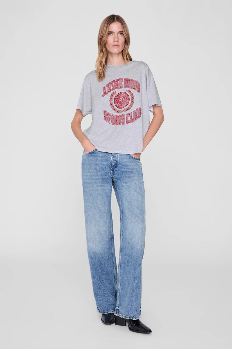 A woman modeling a light gray oversized graphic t-shirt with a distressed red circular logo, paired with light-wash blue jeans and black shoes, against a plain light gray studio background.