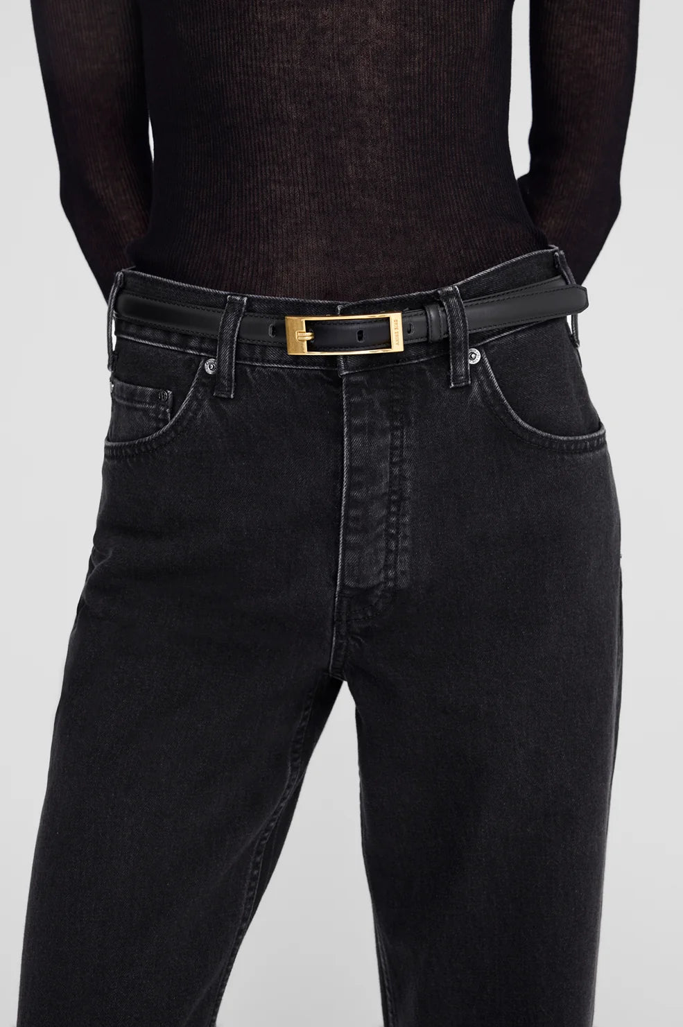 Close-up of a person wearing black jeans with a black belt featuring a gold rectangular buckle, paired with a sheer black long-sleeve top against a light gray background.