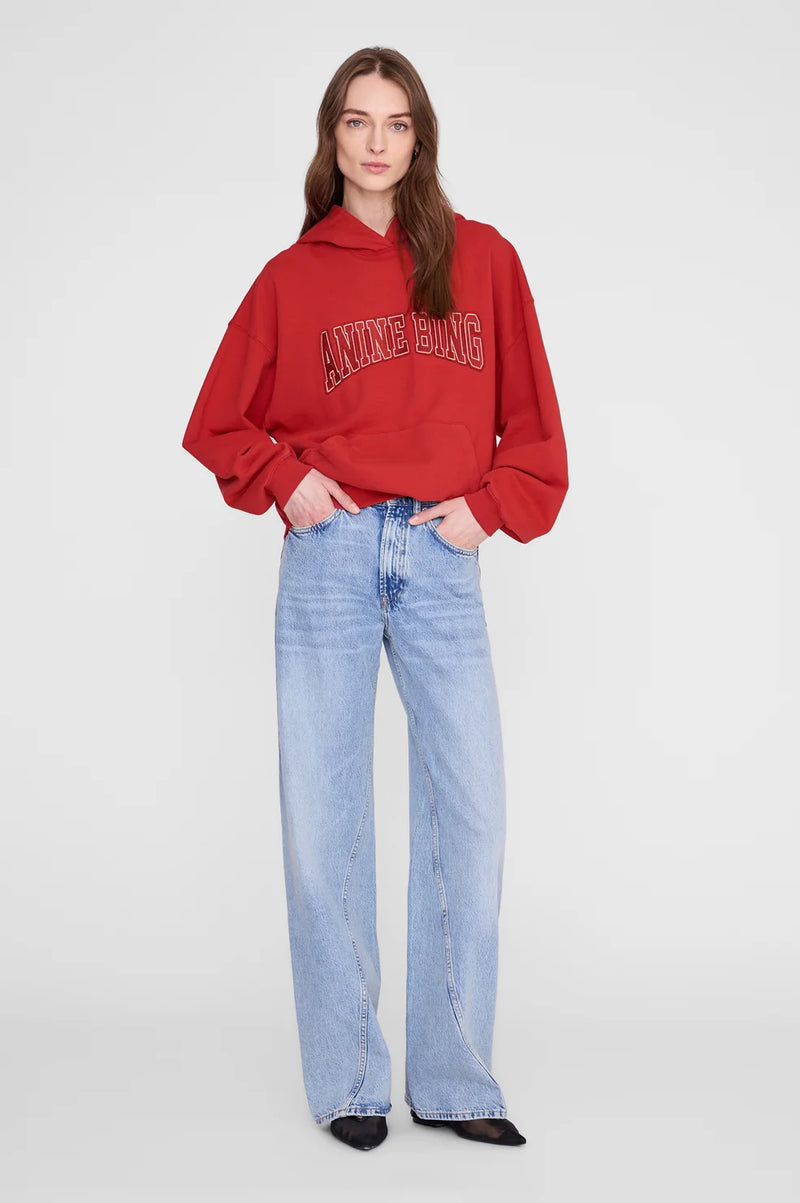 A woman modeling a red cropped hoodie with 'ANNING BANG' embroidered across the chest, paired with light-wash wide-leg jeans and black pointed shoes, against a light gray studio background.